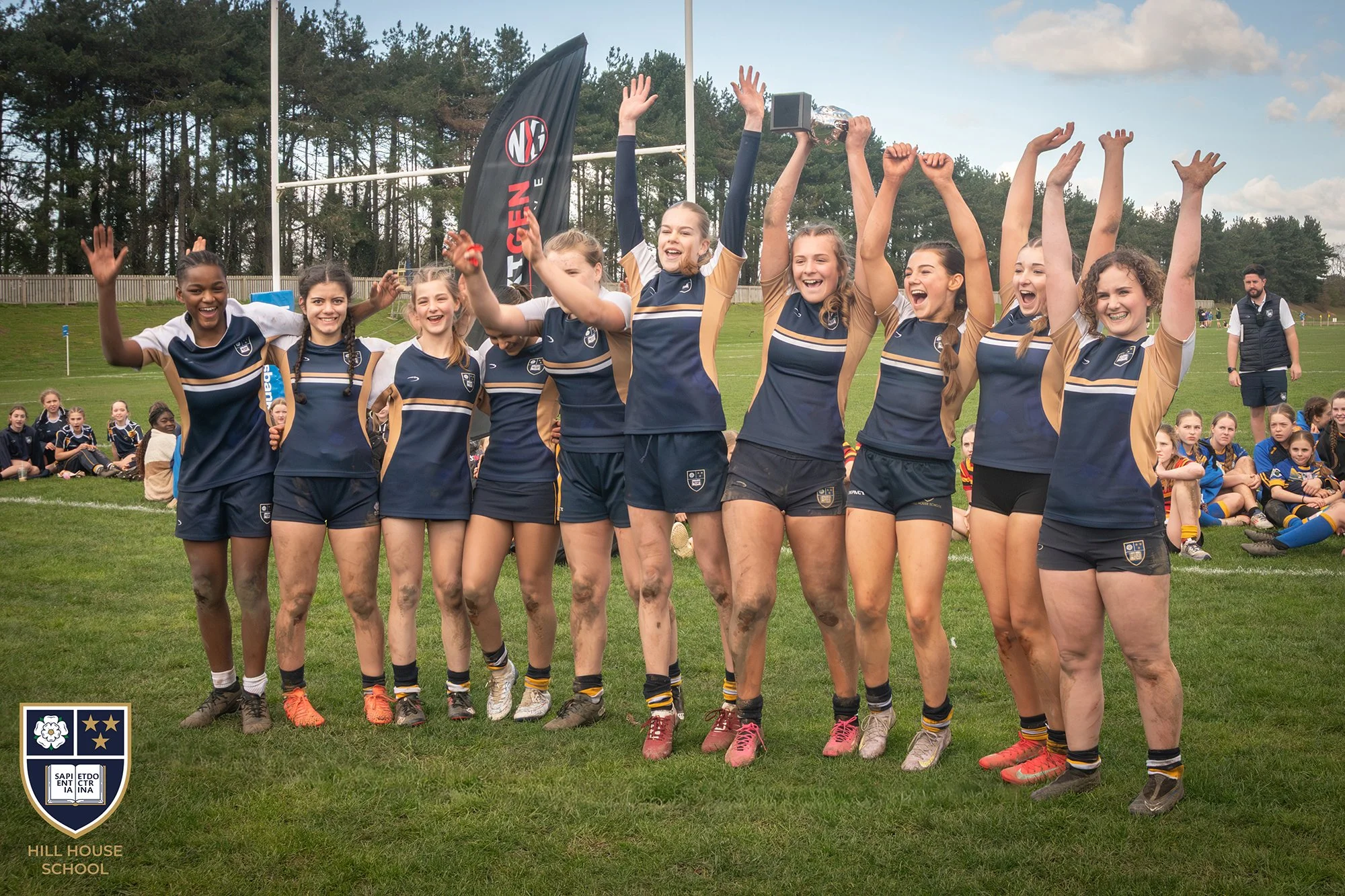 A group of young female rugby players celebrating on a grassy field, some with muddy legs, one holding a trophy, and others clapping and raising their hands. They are wearing matching navy and tan uniforms. In the background, there are other players 