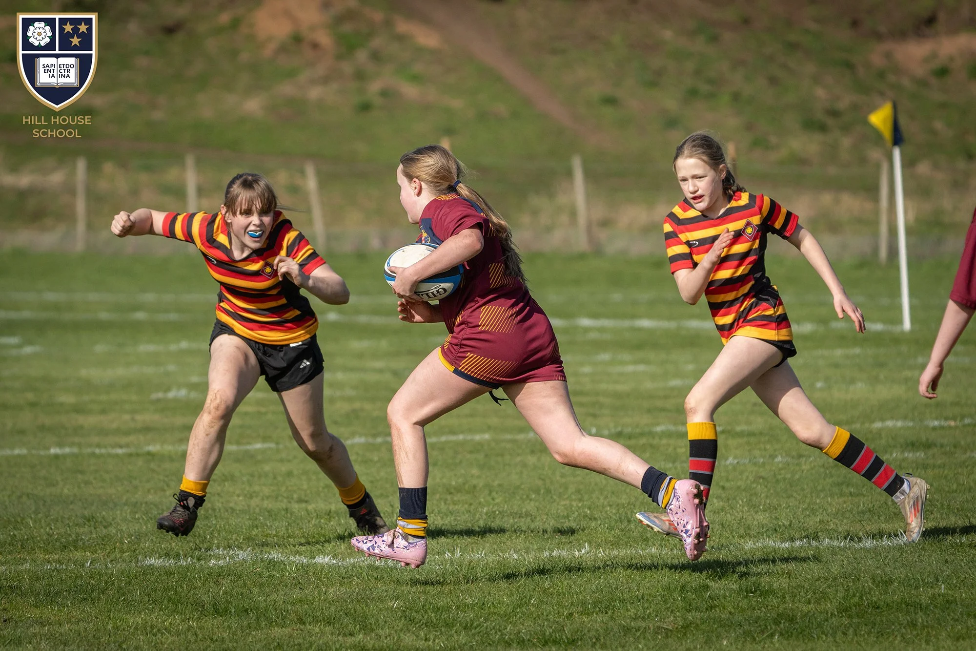 Two young girls playing rugby on a grassy field, wearing red and yellow striped jerseys. One girl is holding a rugby ball and running, while the other girl is chasing her. They appear to be in an intense game.