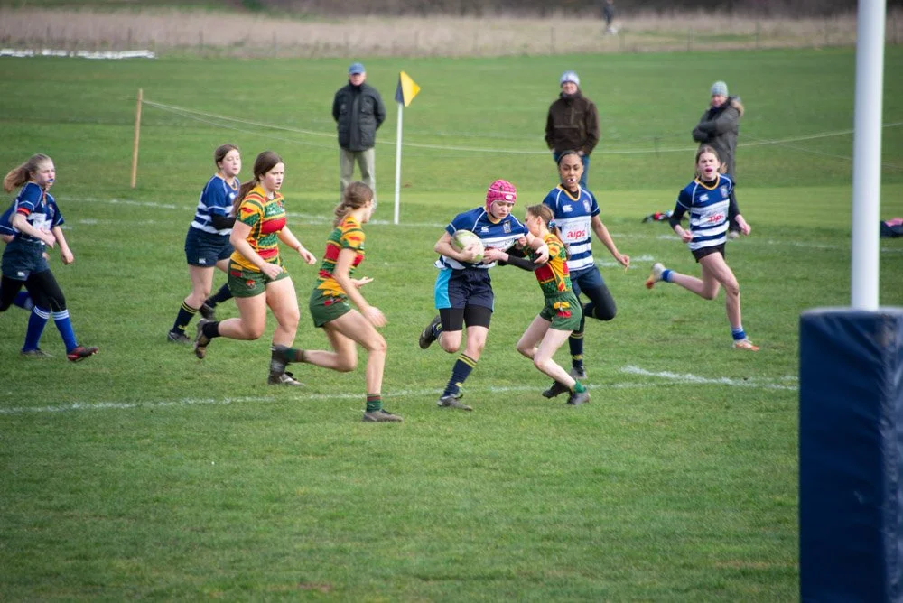 Youth girls playing rugby match on a grassy field, some players in blue and others in colorful striped jerseys, with spectators watching in the background.
