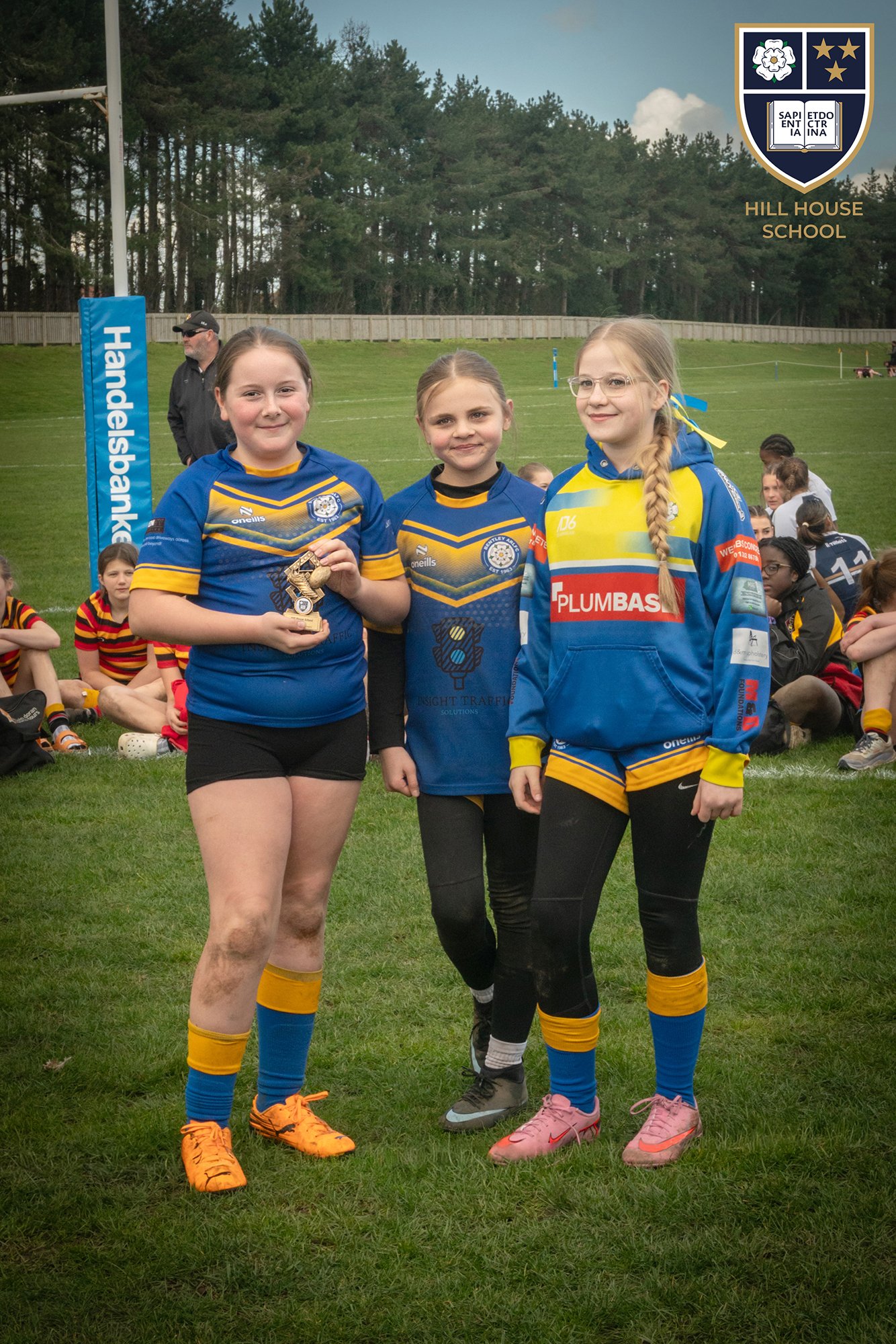 Three young girls in rugby uniforms standing on a field, holding a trophy, with other players and a coach in the background. There is a football goal with a blue padding with the word 'Handelsbank' and a school banner that reads 'Hill House School'.