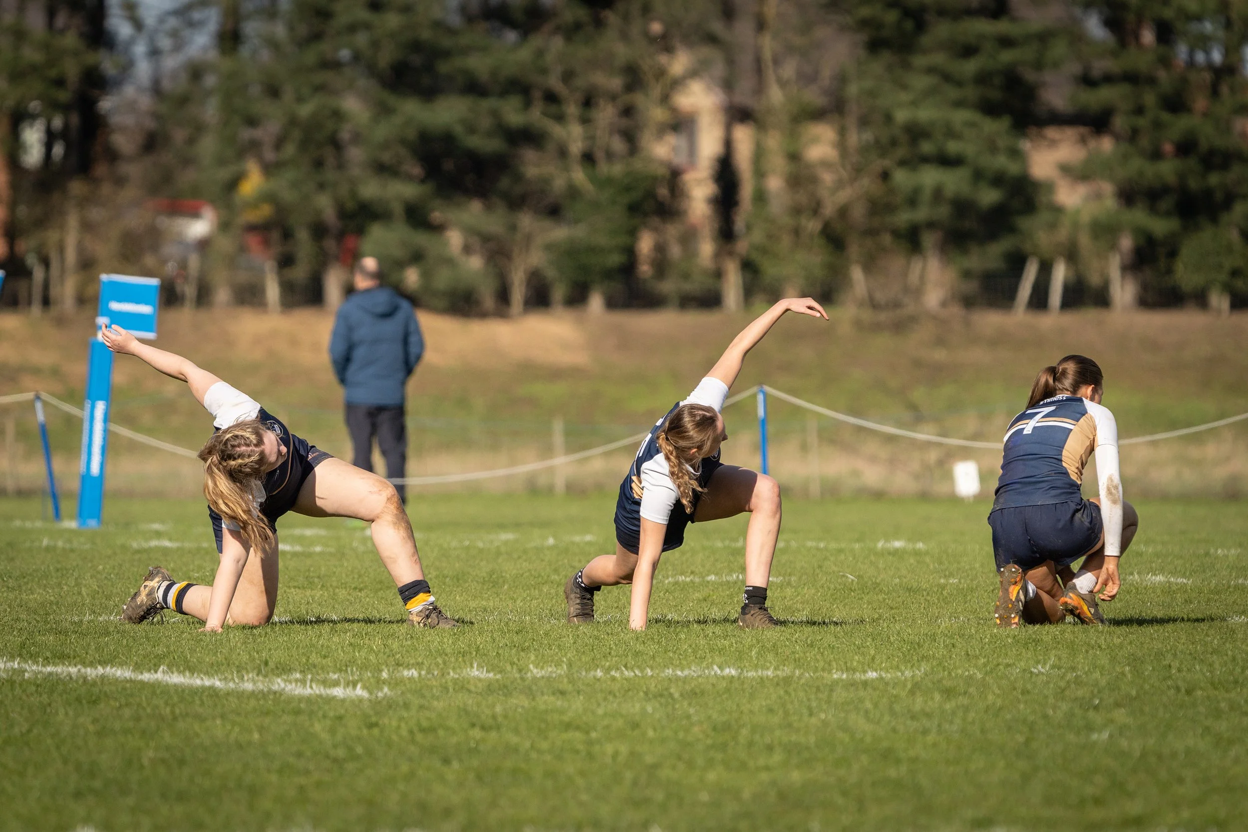 Young women in sports uniforms participating in an outdoor game, kneeling on the grass, with a man watching in the background.