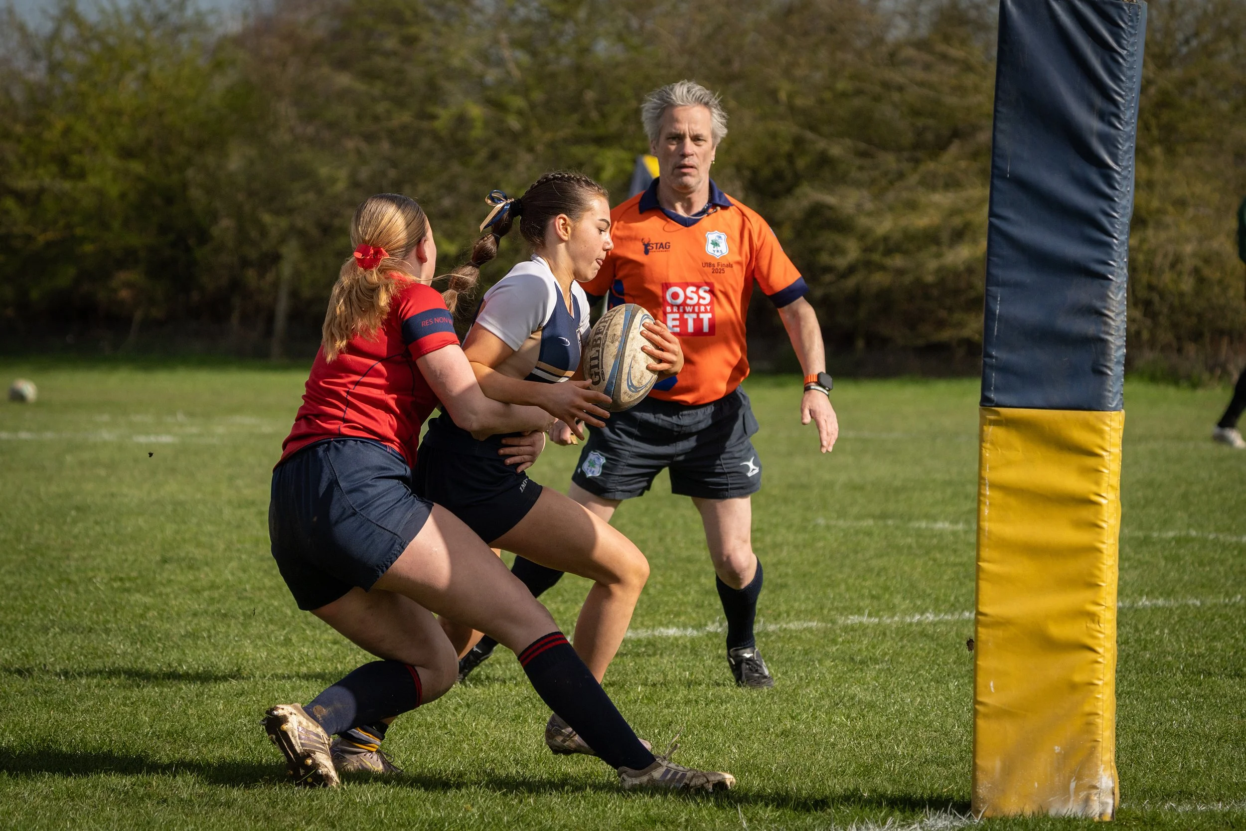 Two female rugby players engaging near a yellow padded goalpost, one is holding a rugby ball, and an official in an orange shirt observing.
