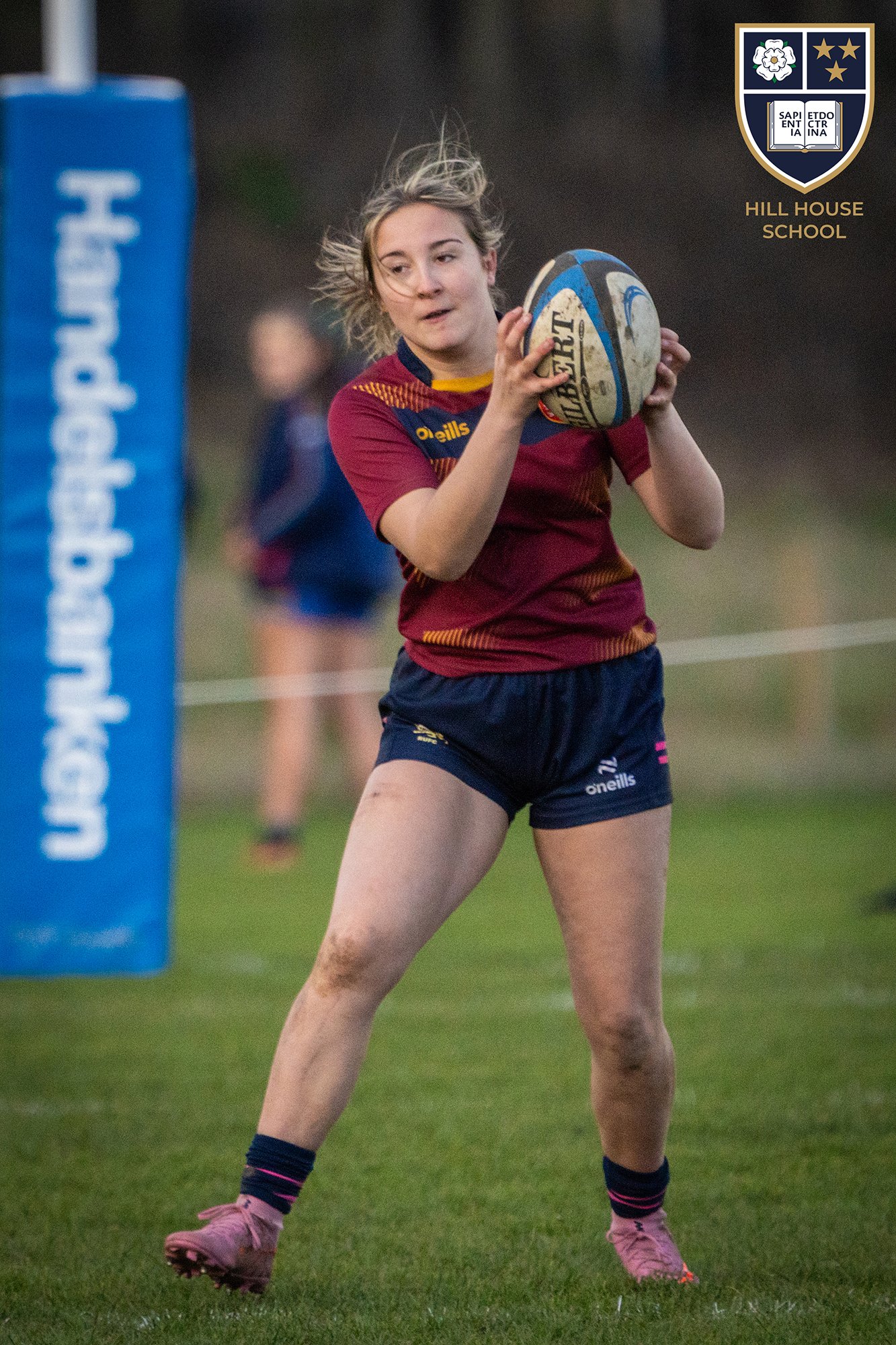 A female rugby player holding a rugby ball during a match, wearing a maroon and navy sports uniform, pink cleats, and navy socks with pink stripes. She has blonde hair and is standing on a grass field with a blurred background. A blue padded post is 