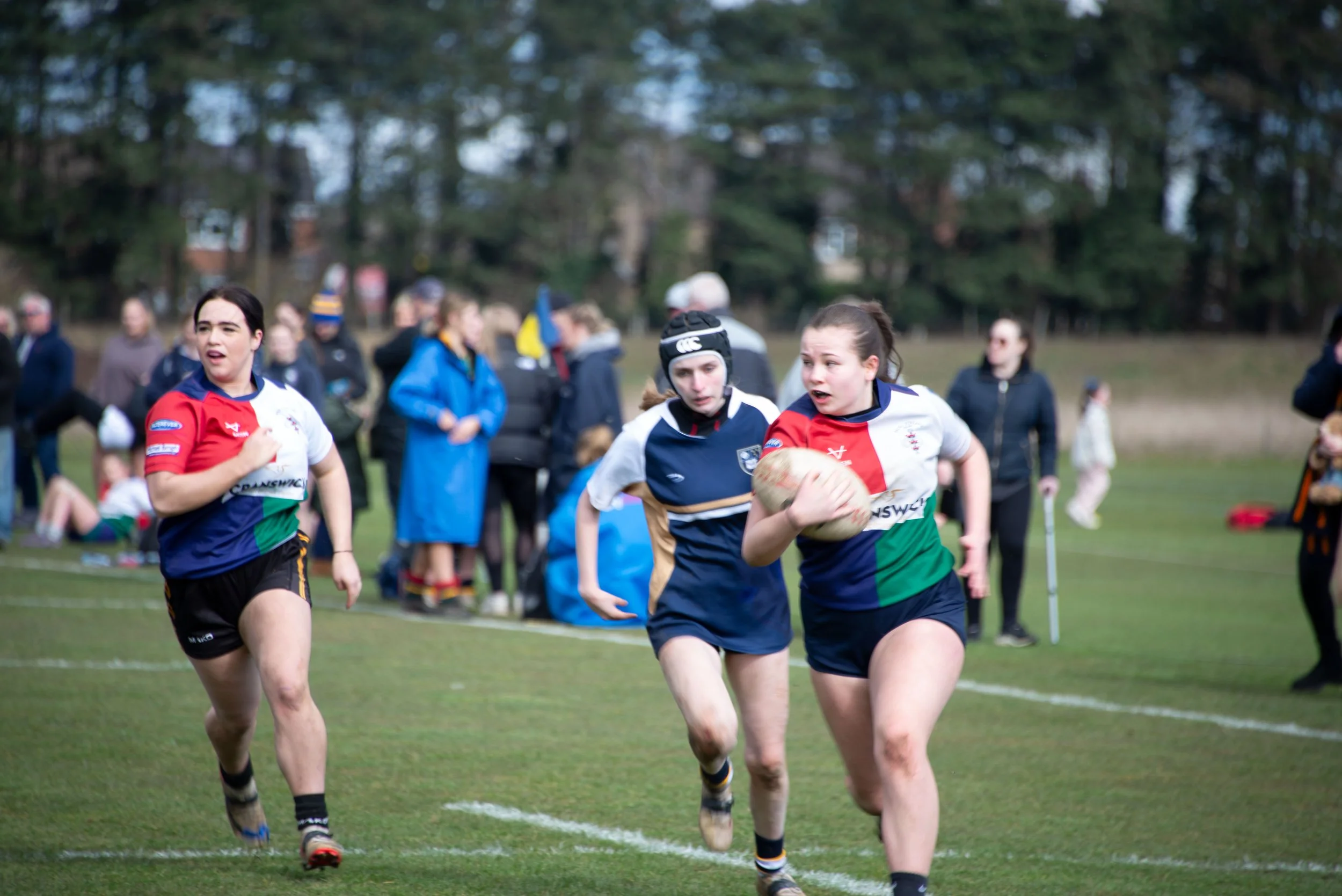 Women playing rugby on a grassy field with spectators in the background.