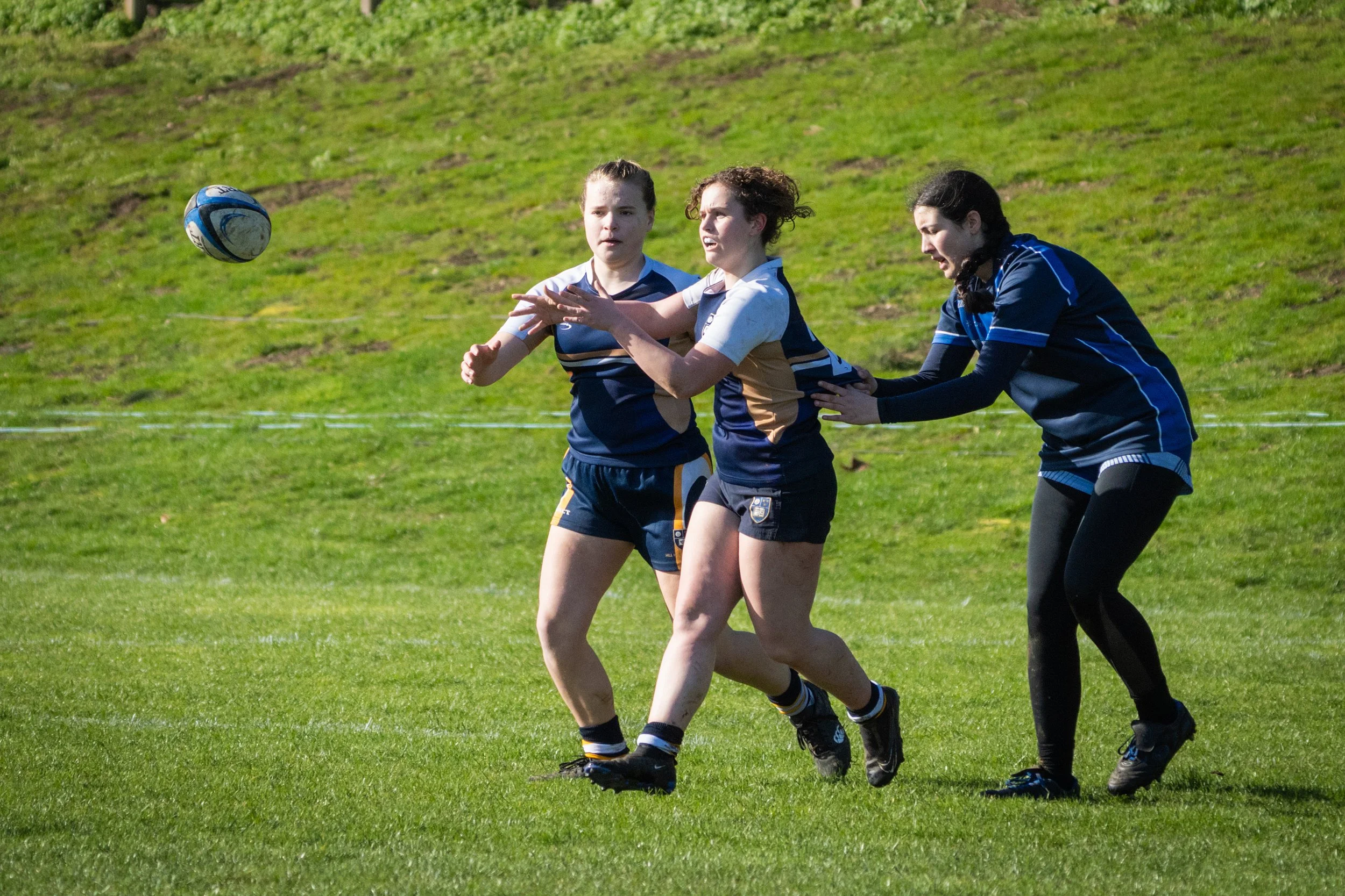 Three female rugby players wearing blue and black uniforms play on a green field, with one of them reaching towards a rugby ball in midair.