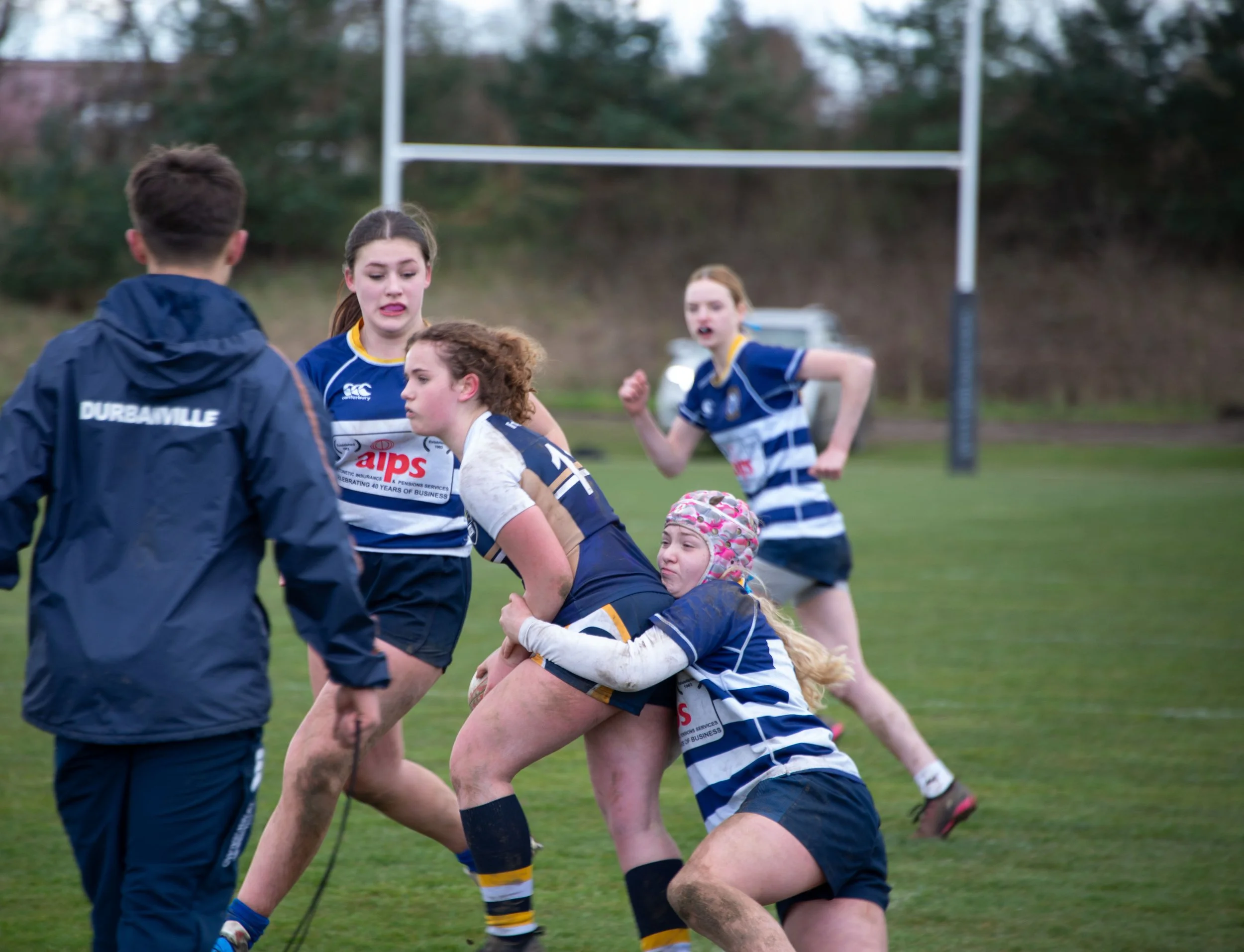 Rugby players engaged in a tackle on a grassy field with goalposts in the background.