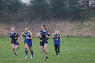 Four young girls running on a grassy field, wearing matching blue and yellow sports uniforms.