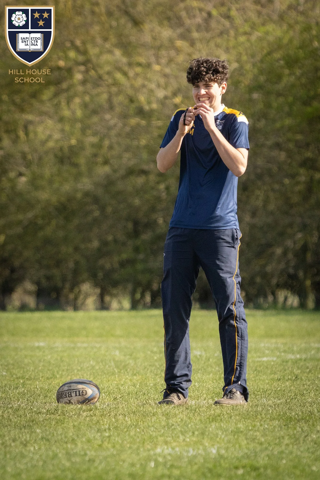 A young man in a navy and yellow sports uniform standing on a grass field, smiling with teeth visible, with a rugby ball on the ground nearby and trees in the background.