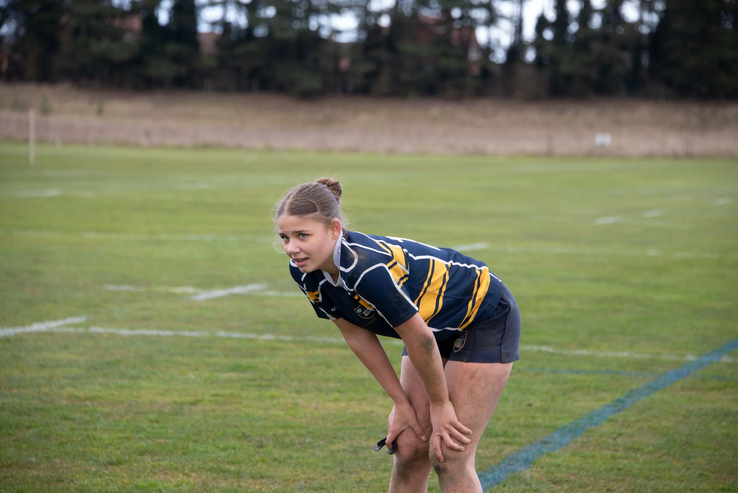 A female soccer player crouched down on a grassy field, wearing a navy and yellow uniform, with a focused expression.