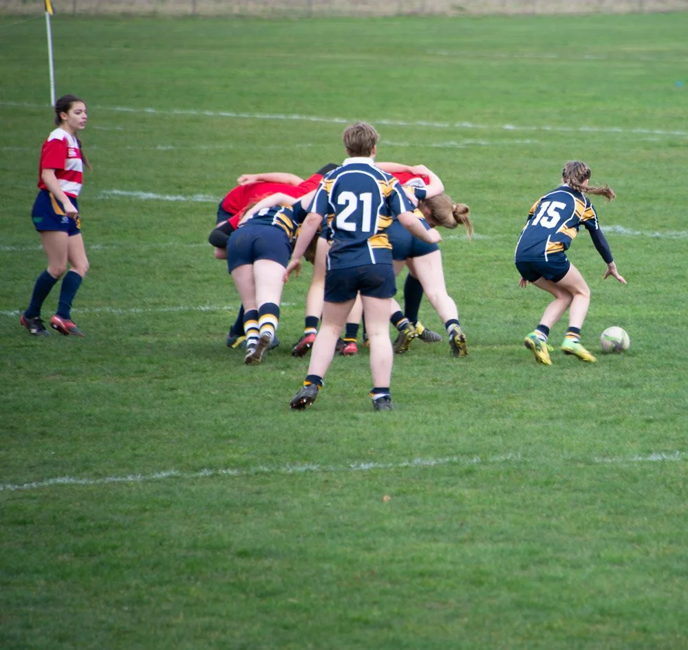 Girls playing rugby on a grass field, with one girl in a navy and yellow uniform preparing to kick the rugby ball.