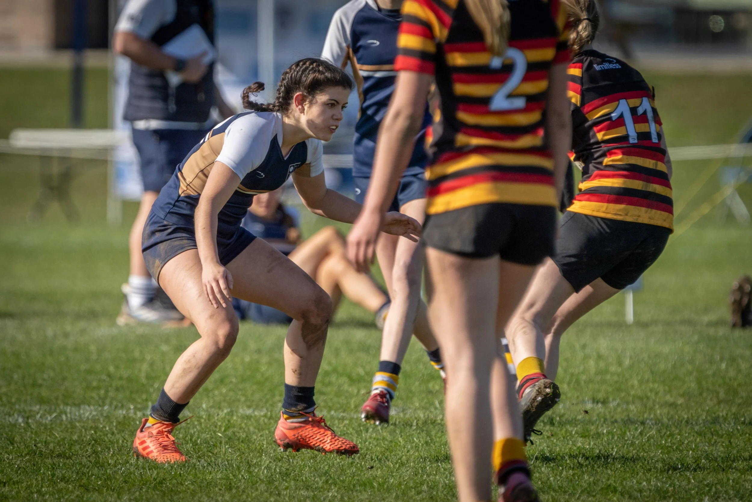 Women's rugby match on a grassy field with players in navy, white, and striped uniforms, some with muddy legs, and orange and red cleats.