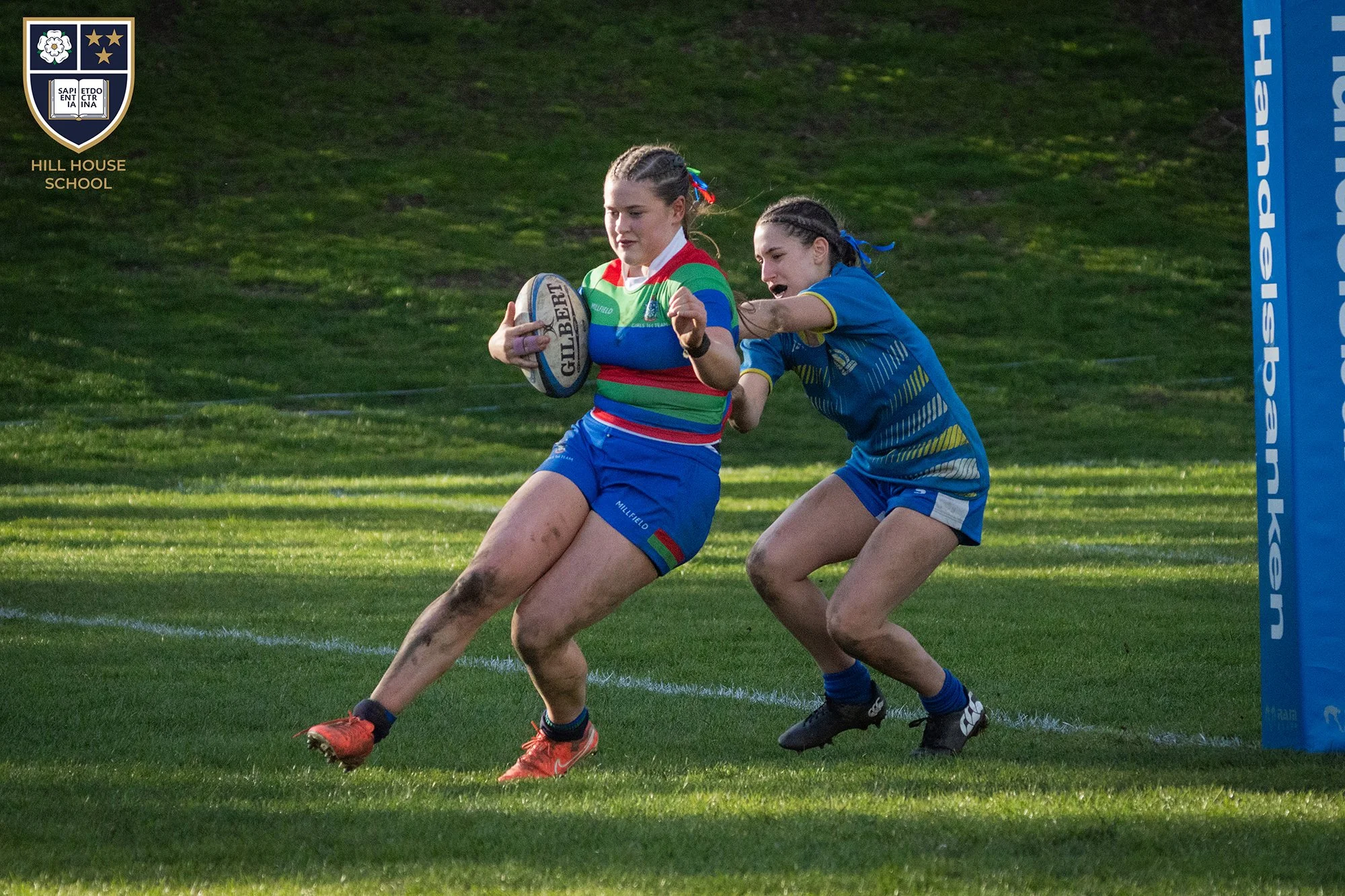 Two female rugby players are competing for the ball near a try line on a grassy field. The player in a red, green, and blue striped jersey holds the ball, while the other in a blue jersey reaches to block her. A blue padded post is visible on the rig