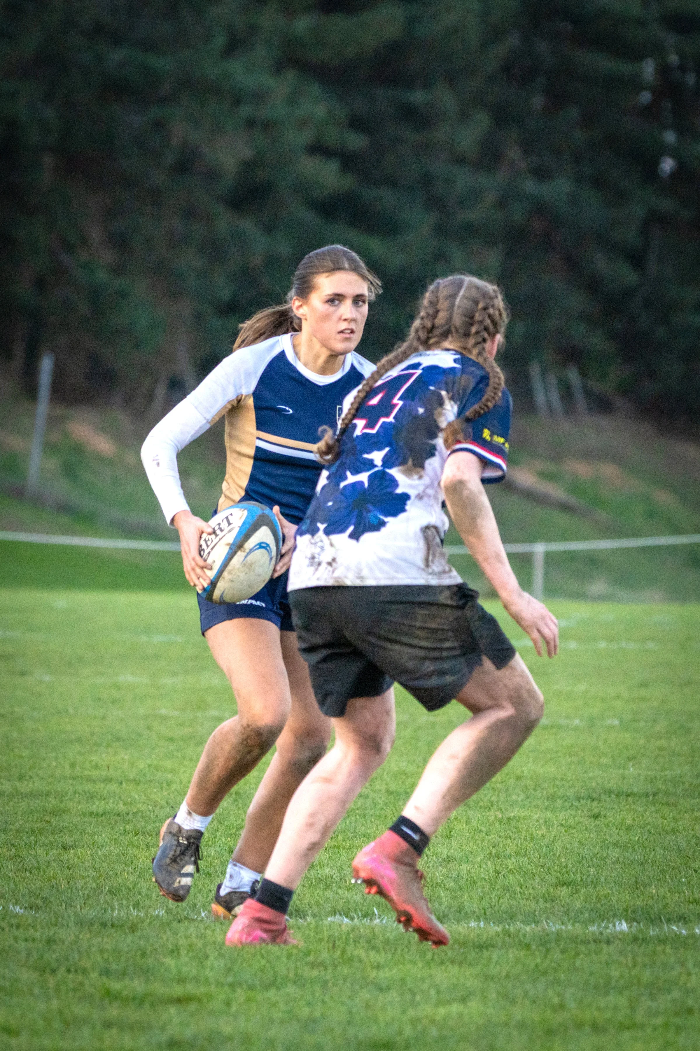 Two women playing rugby outdoors on a grassy field, with a background of trees. One woman is holding a rugby ball and wearing a blue and white jersey, while the other woman, with braided hair, is about to tackle or block her.