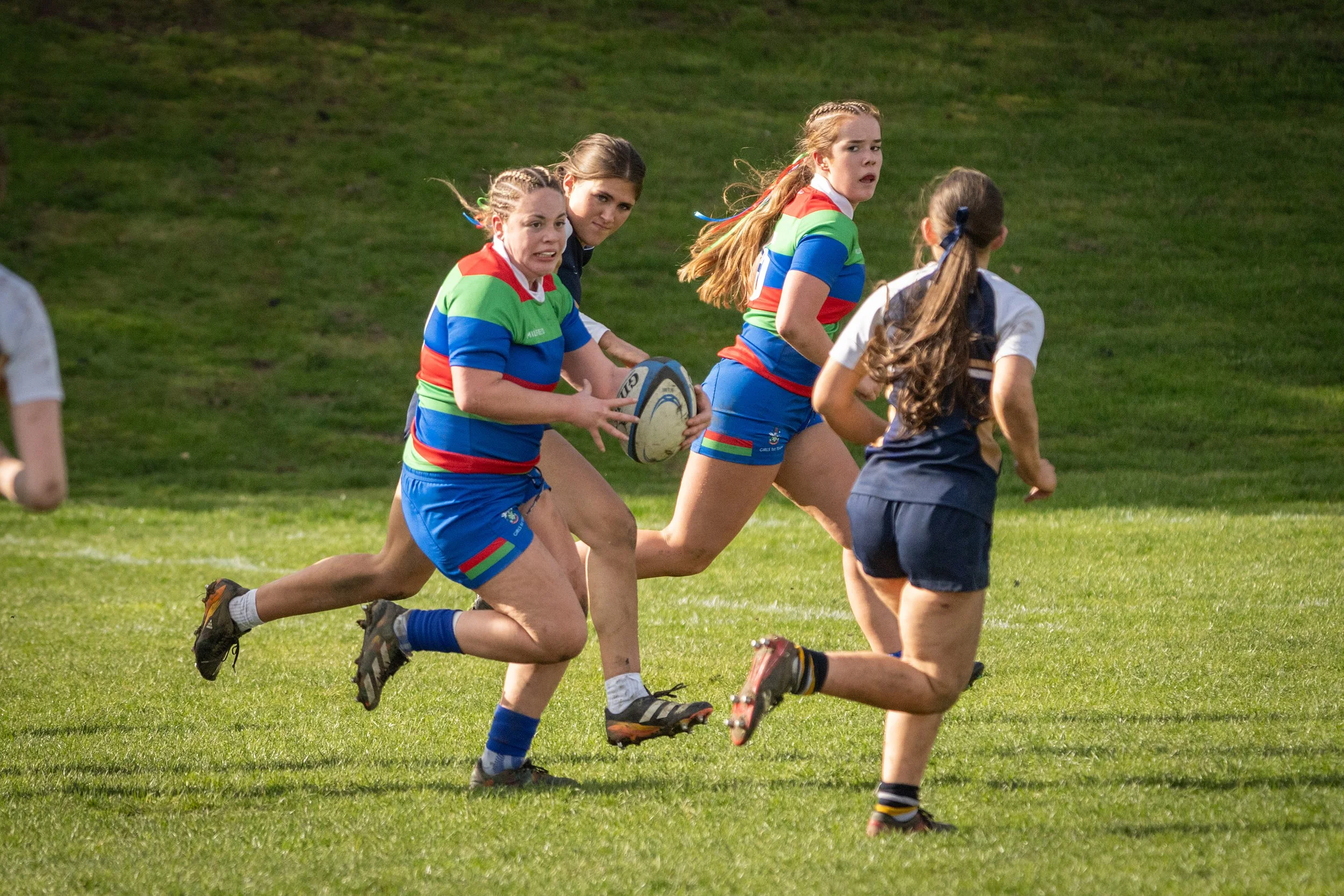 Women in colorful rugby uniforms running on a grassy field during a match.
