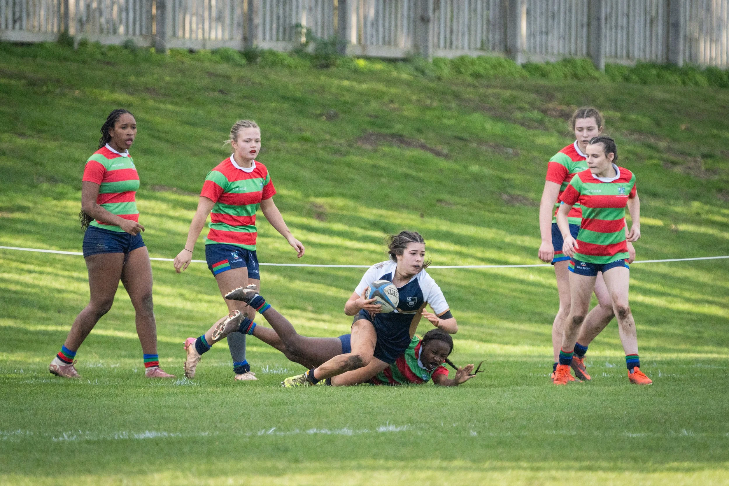 Girls playing rugby on a grassy field, with some girls in red and green striped jerseys and a player in black with a rugby ball