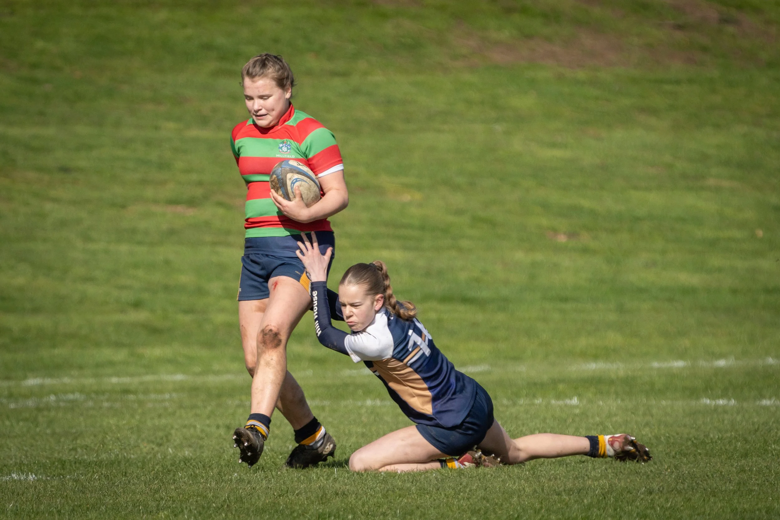 A female rugby player in a green and red striped jersey holding a rugby ball while standing on a grassy field, with a player in a blue and yellow uniform on the ground reaching out to her.