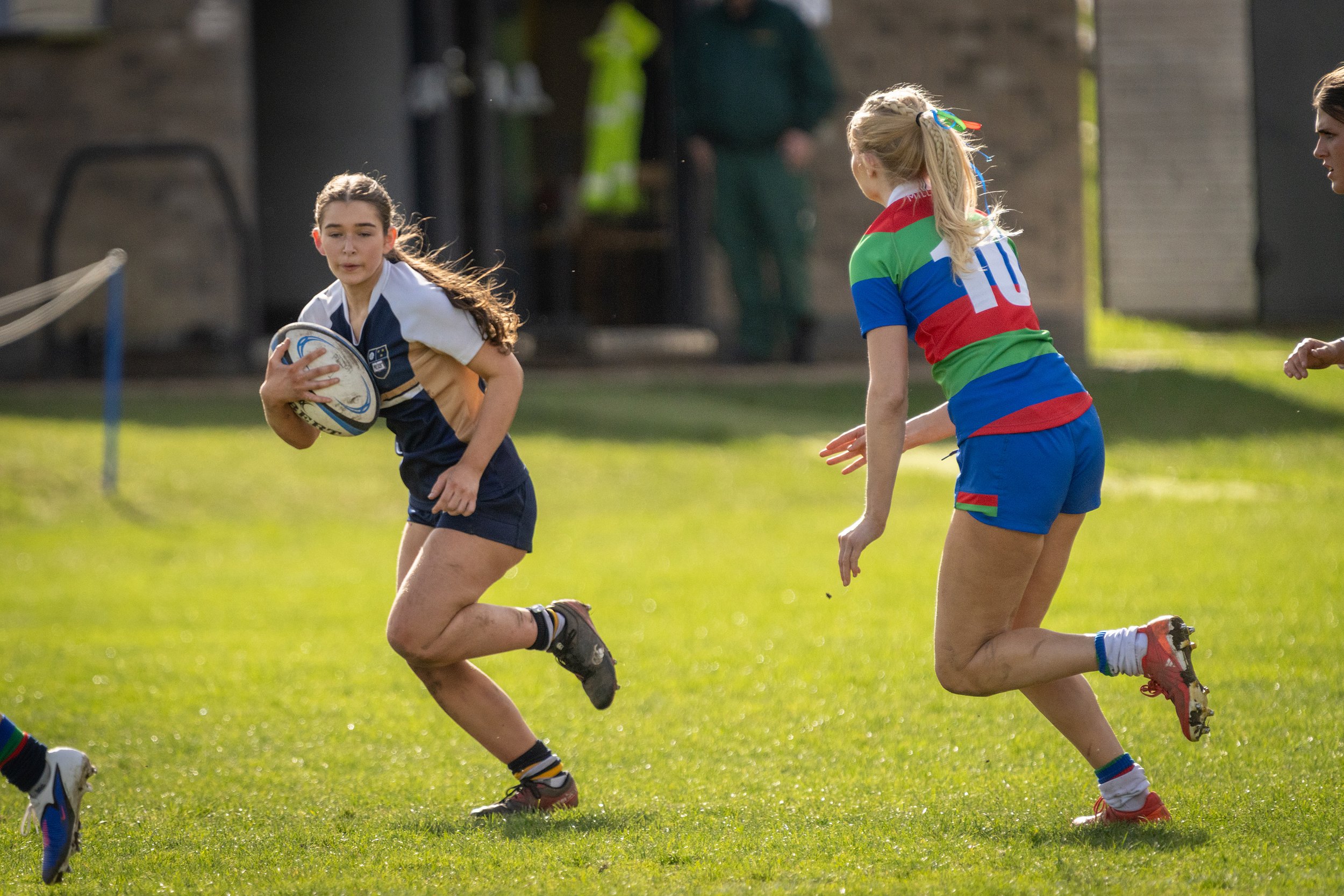 Girls playing rugby on a grassy field, with one girl carrying the ball and others running nearby.