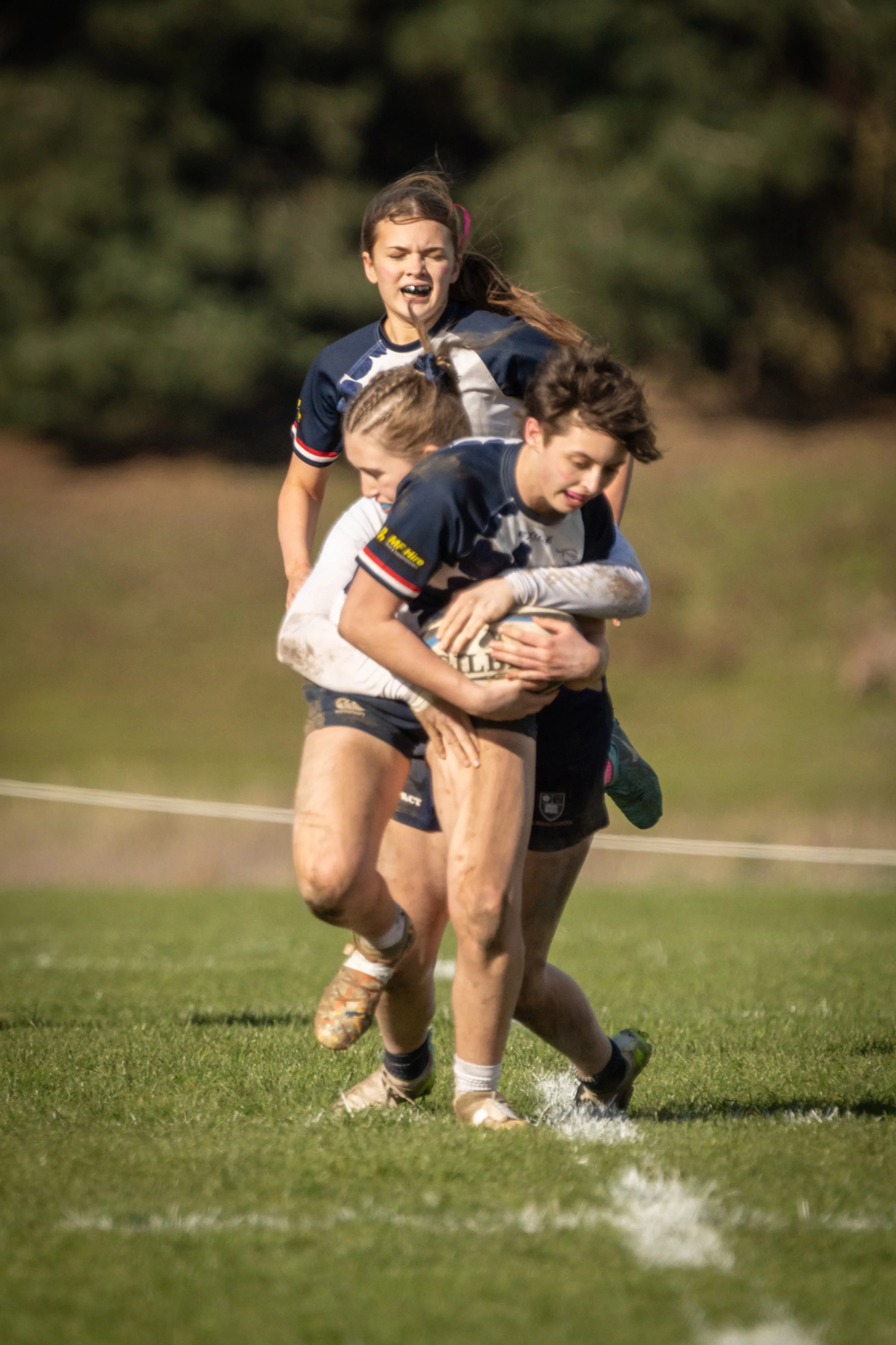Two rugby players, a girl and a boy, are competing for possession of a rugby ball on a grassy field. The girl is wearing a navy blue and white jersey with her hair tied back, and appears to be distressed or exerting effort. The boy, also in a the nav