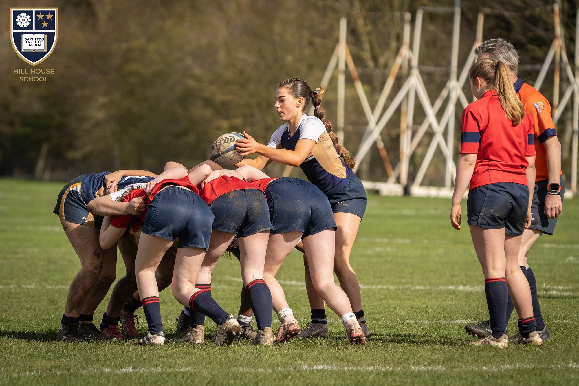 Rugby team preparing for a scrum on a grassy field, with coach supervising. Players are dressed in navy, red, and orange jerseys, some with muddy shorts.