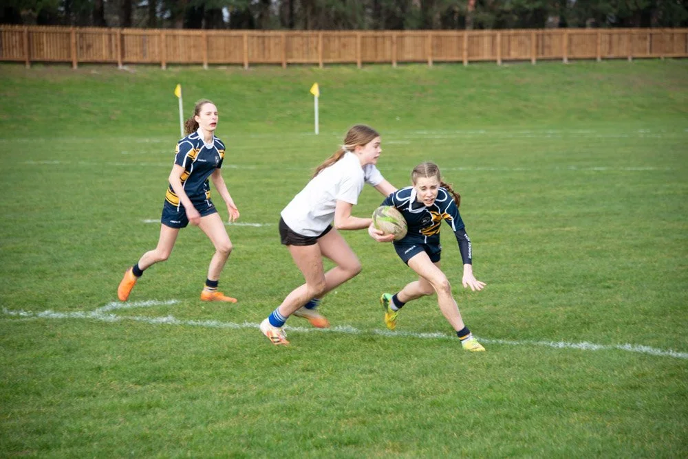 Girls playing rugby on a grassy field, with one girl running while holding a rugby ball, and two other girls attempting to stop her.