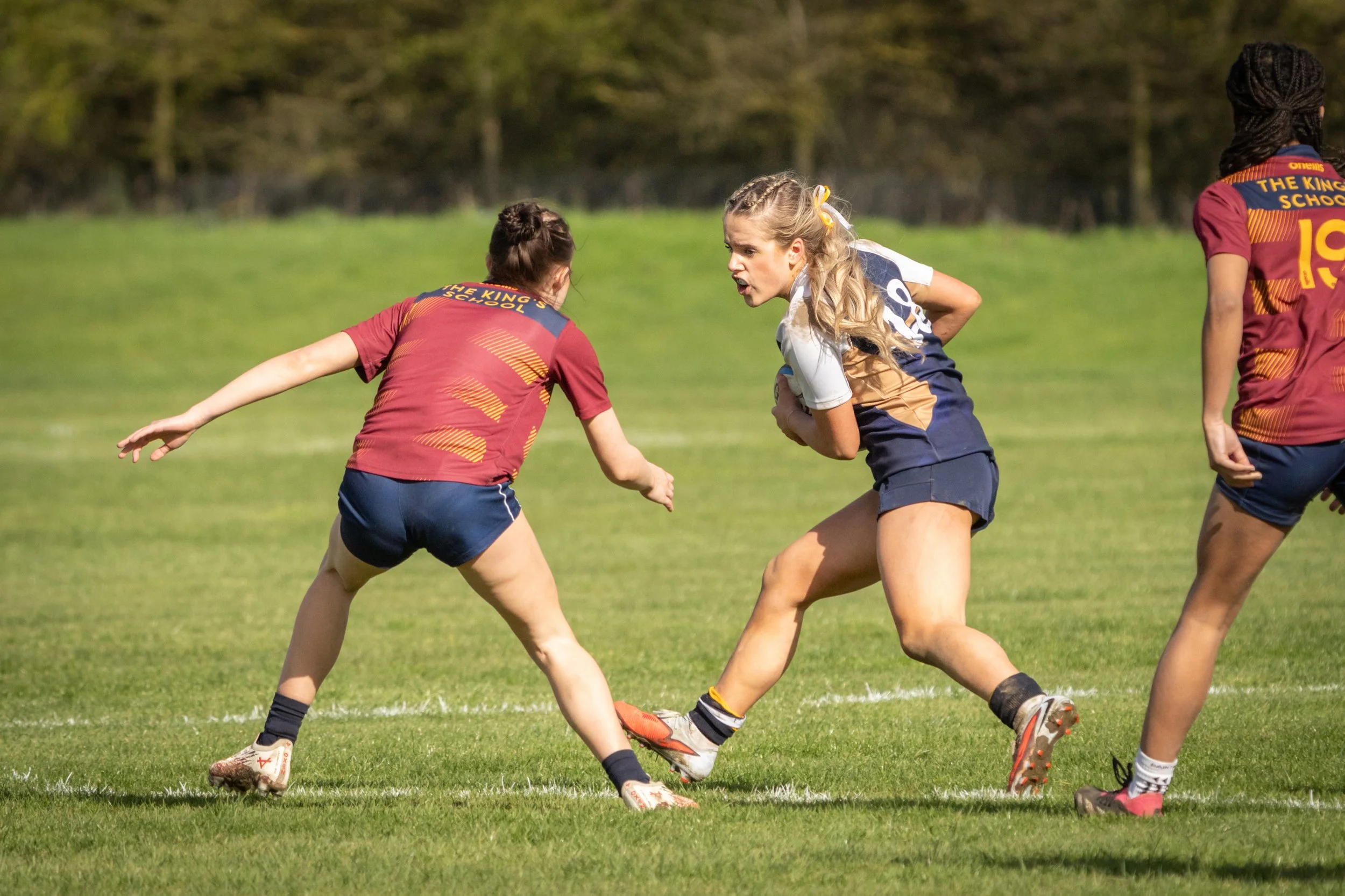 Girls playing rugby on a grassy field with trees in the background.