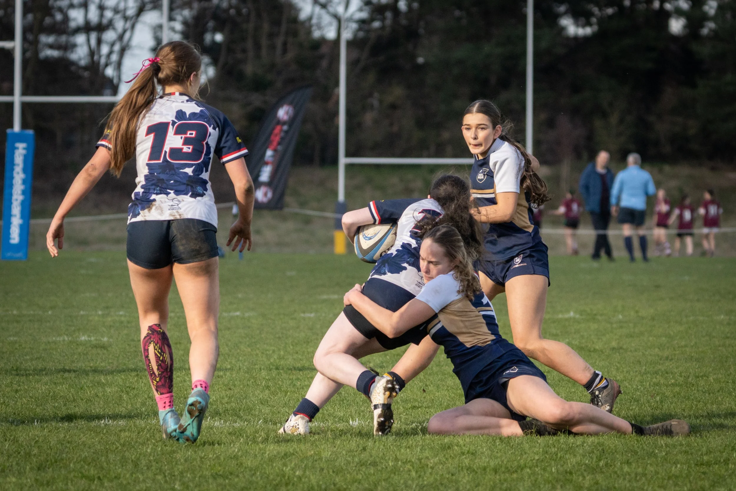 Women playing rugby on a grass field, some women are standing while others are on the ground, with one holding a rugby ball. In the background, there are trees and a few spectators.