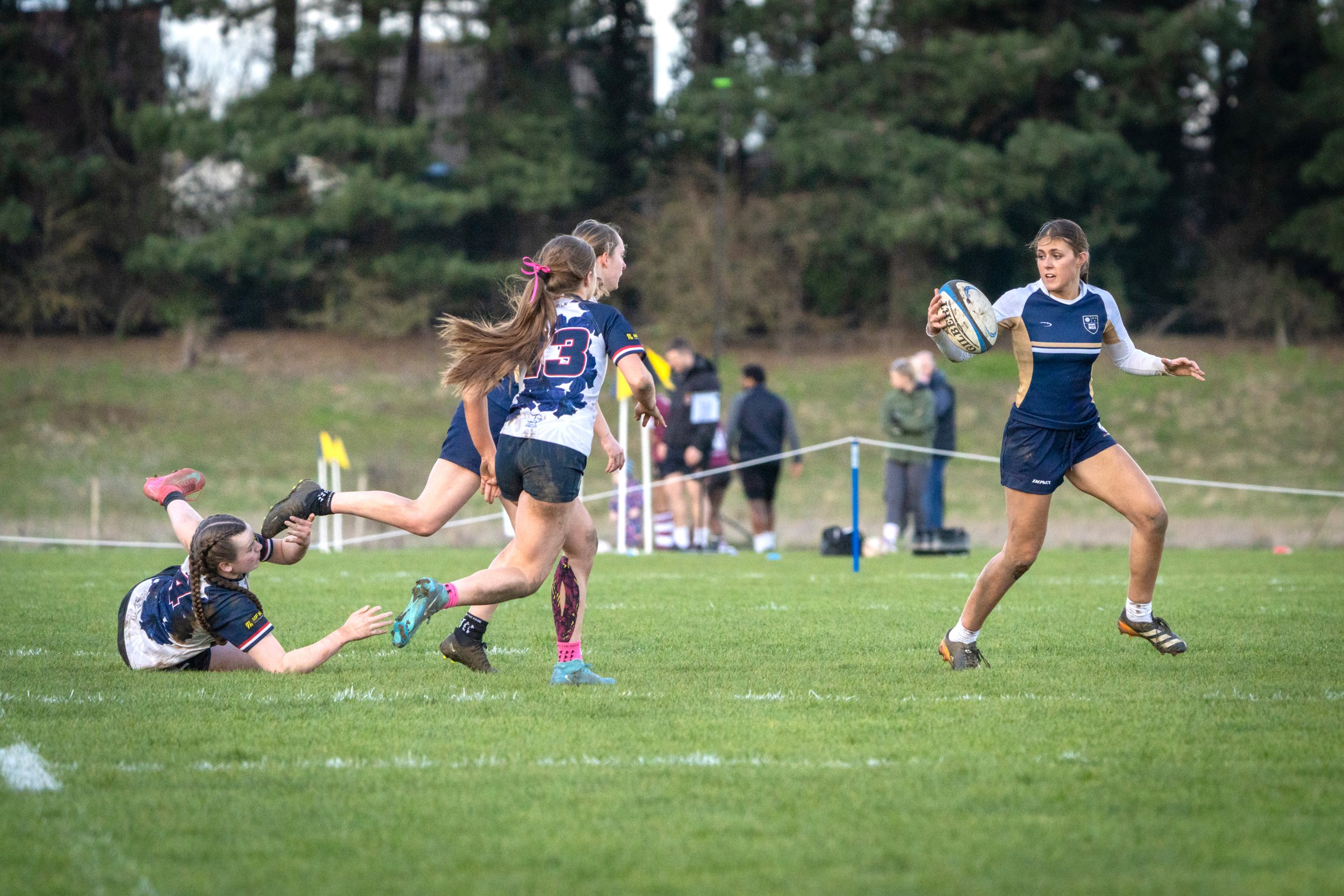 Girls playing rugby outdoors on the field during daytime, with trees in the background.