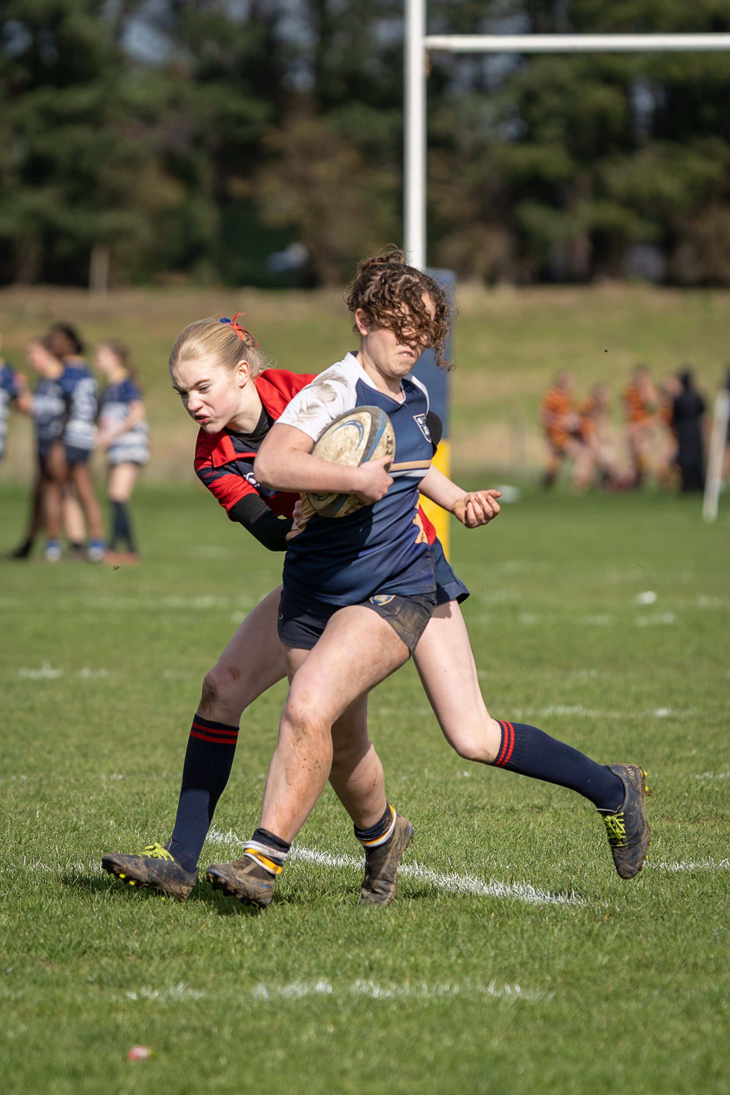Two young women playing rugby on a grassy field during daytime, with one holding a rugby ball while the other attempts to tackle her. In the background, other players and trees are visible.