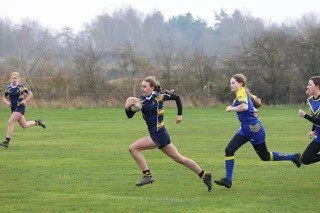 Girls playing rugby on a grassy field, running with a rugby ball.
