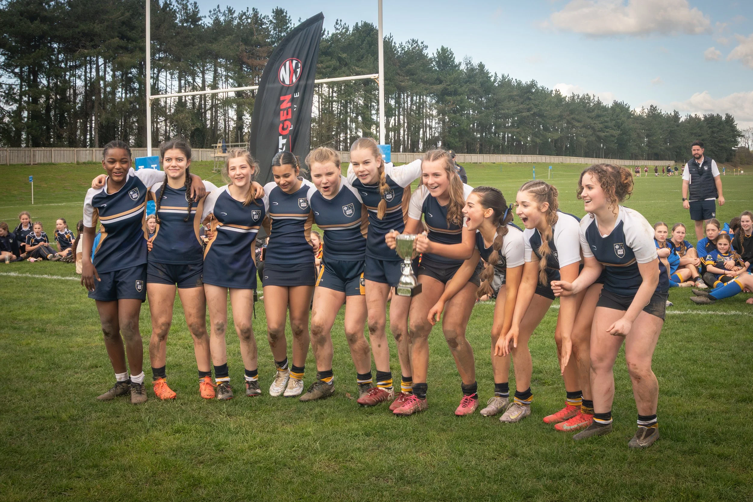 A group of young female rugby players celebrating and smiling on a grassy field, with a trophy in front of them and other players seated behind them, against a backdrop of trees and a flag.