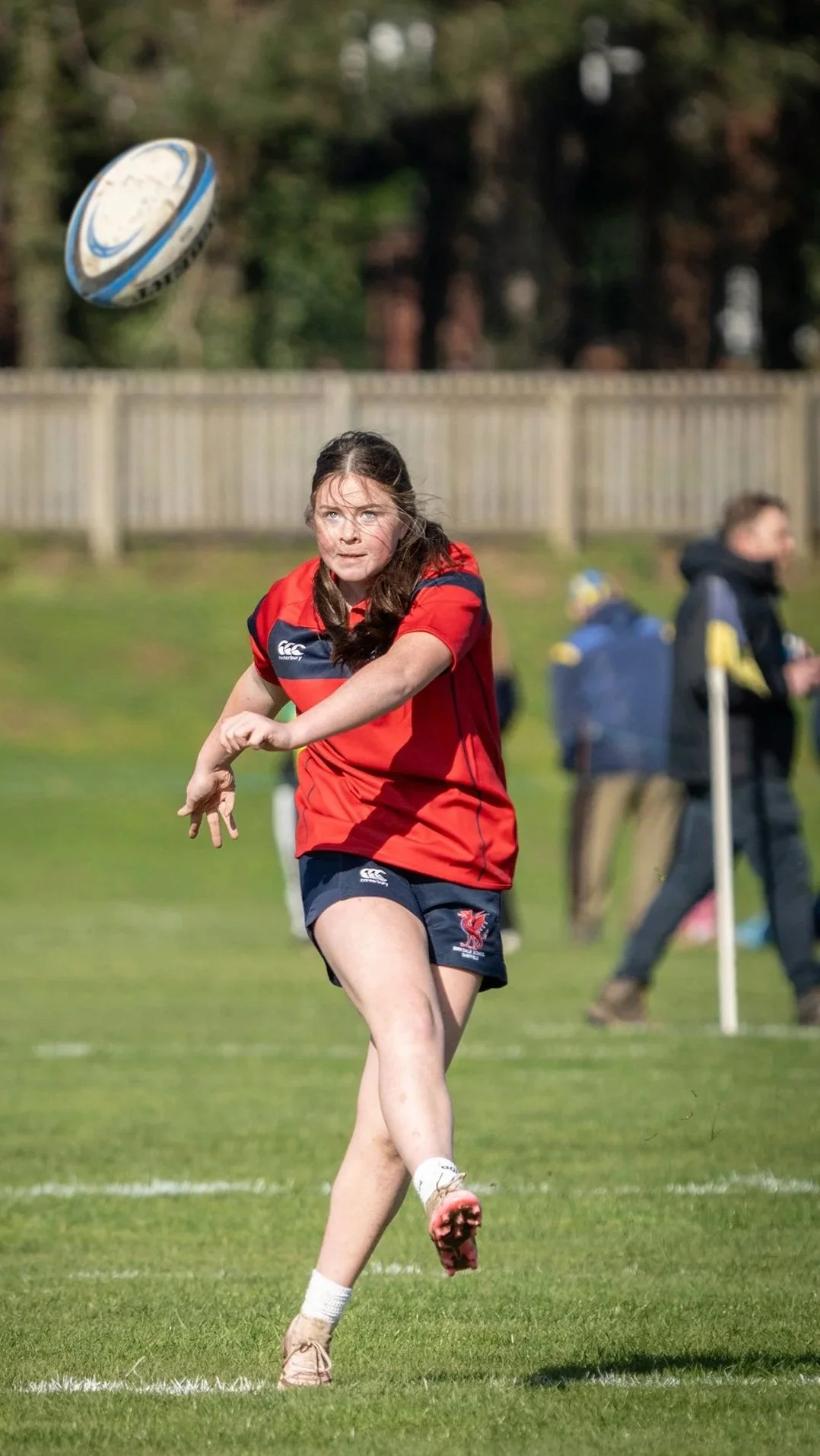 A female rugby player in a red and blue uniform is running on a grassy field, about to catch a rugby ball in mid-air. In the background, a fence and a person walking are visible.