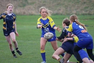Girls playing rugby on a grassy field, with one girl passing the ball as others defend.