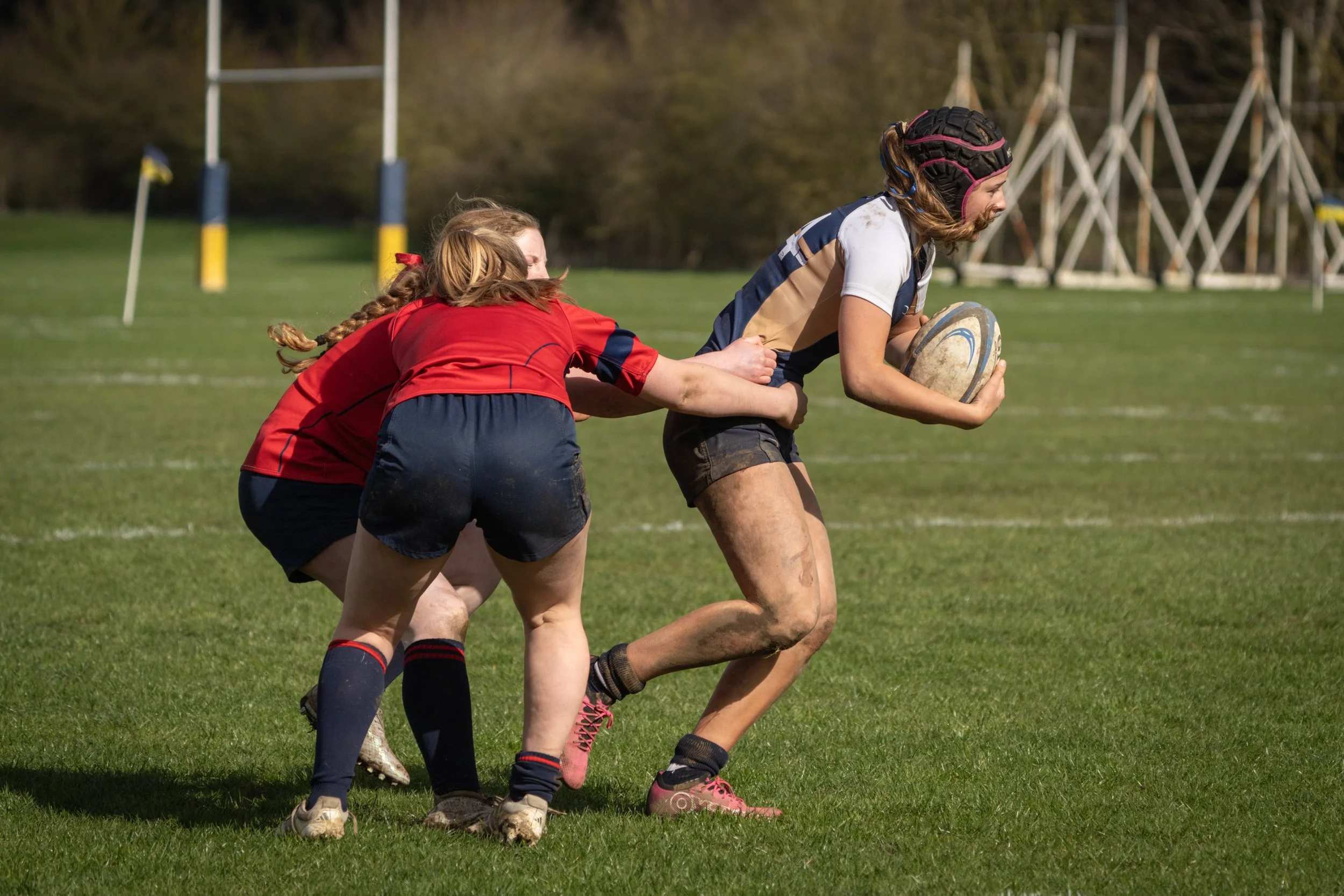 Girls playing rugby on a grassy field, one girl running with a rugby ball, other girls trying to grasp her from behind, all wearing sports uniforms with muddy knees and elbows.