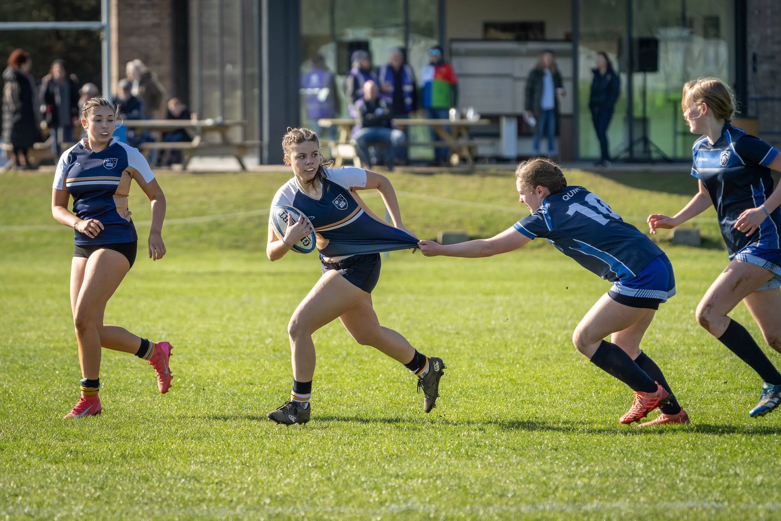 Women playing rugby on a grassy field during daytime with spectators in the background.