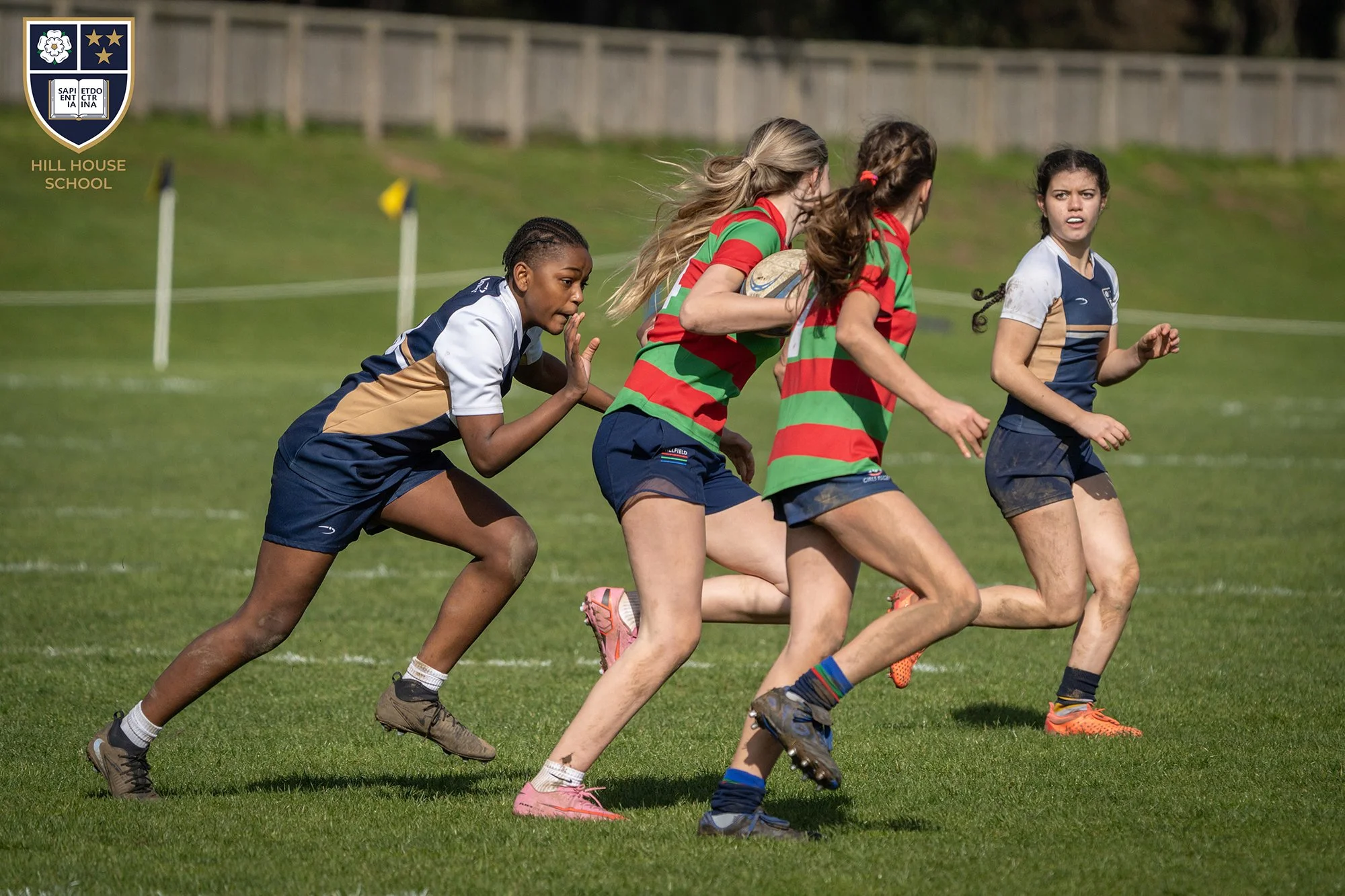 Girls playing rugby on a grassy field, wearing sports uniforms with one girl holding a rugby ball as they run.