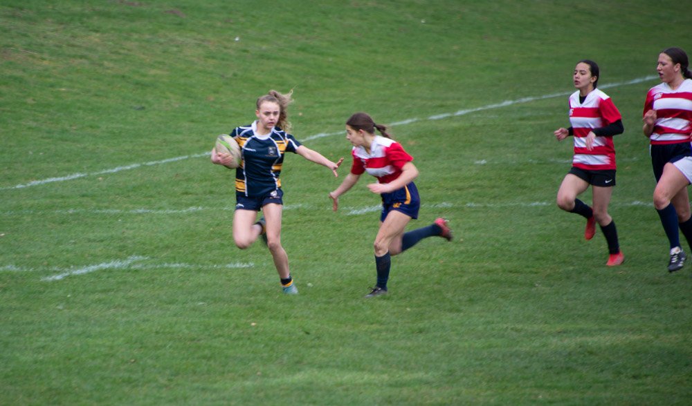 Girls playing rugby on a grassy field, some wearing red and white striped jerseys, one girl with a blue and yellow jersey holding the ball, other girls chasing after her.