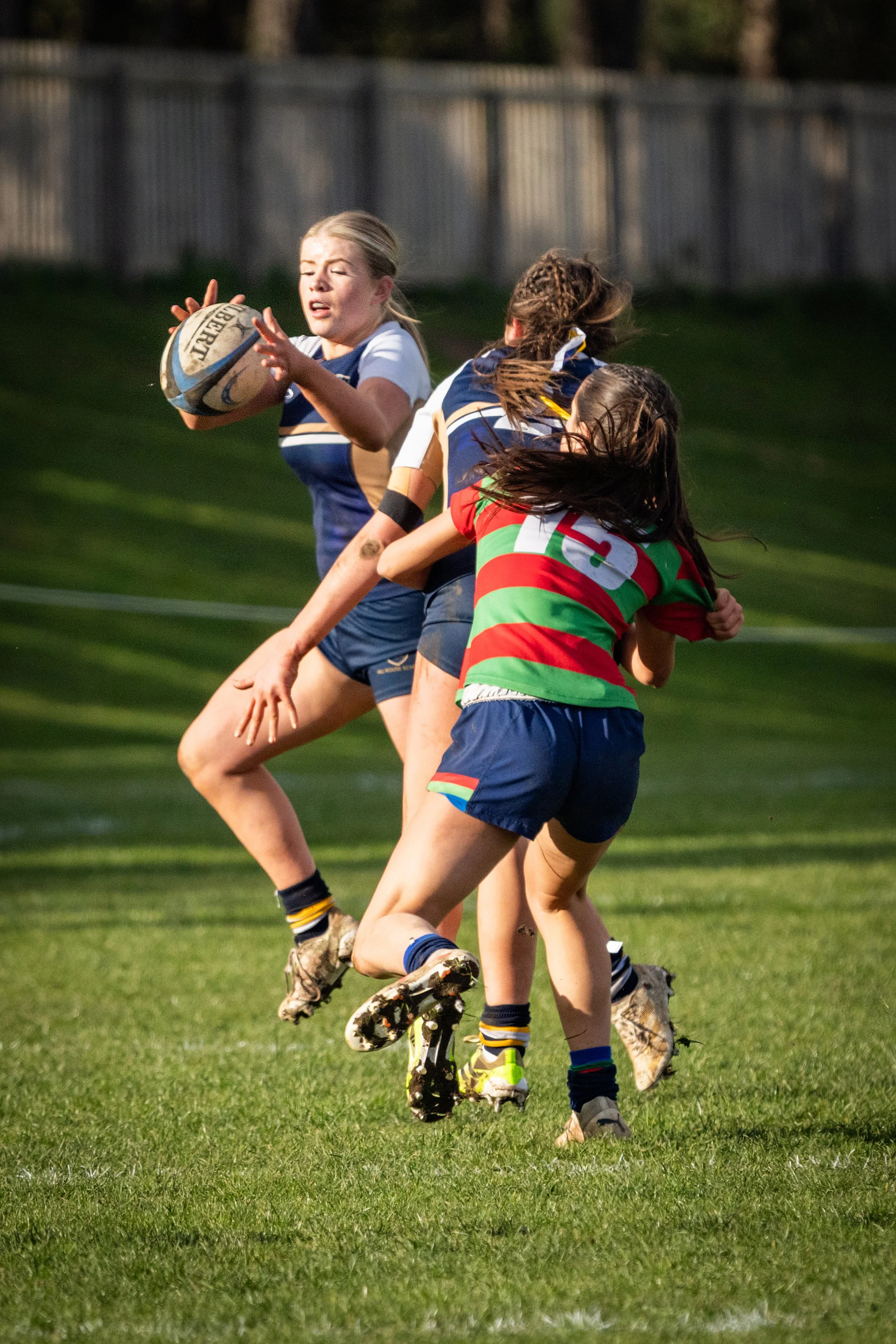 Women playing rugby on a grassy field, with one woman holding a rugby ball and others tackling or chasing her, all wearing sports uniforms.