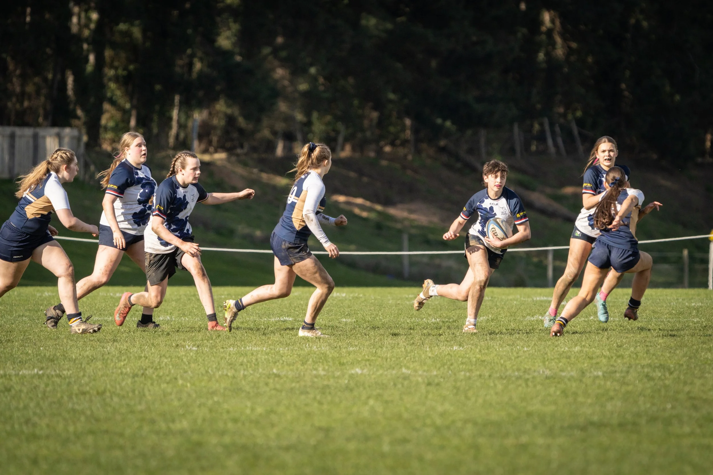 Young girls playing rugby on a grassy field with trees in the background.