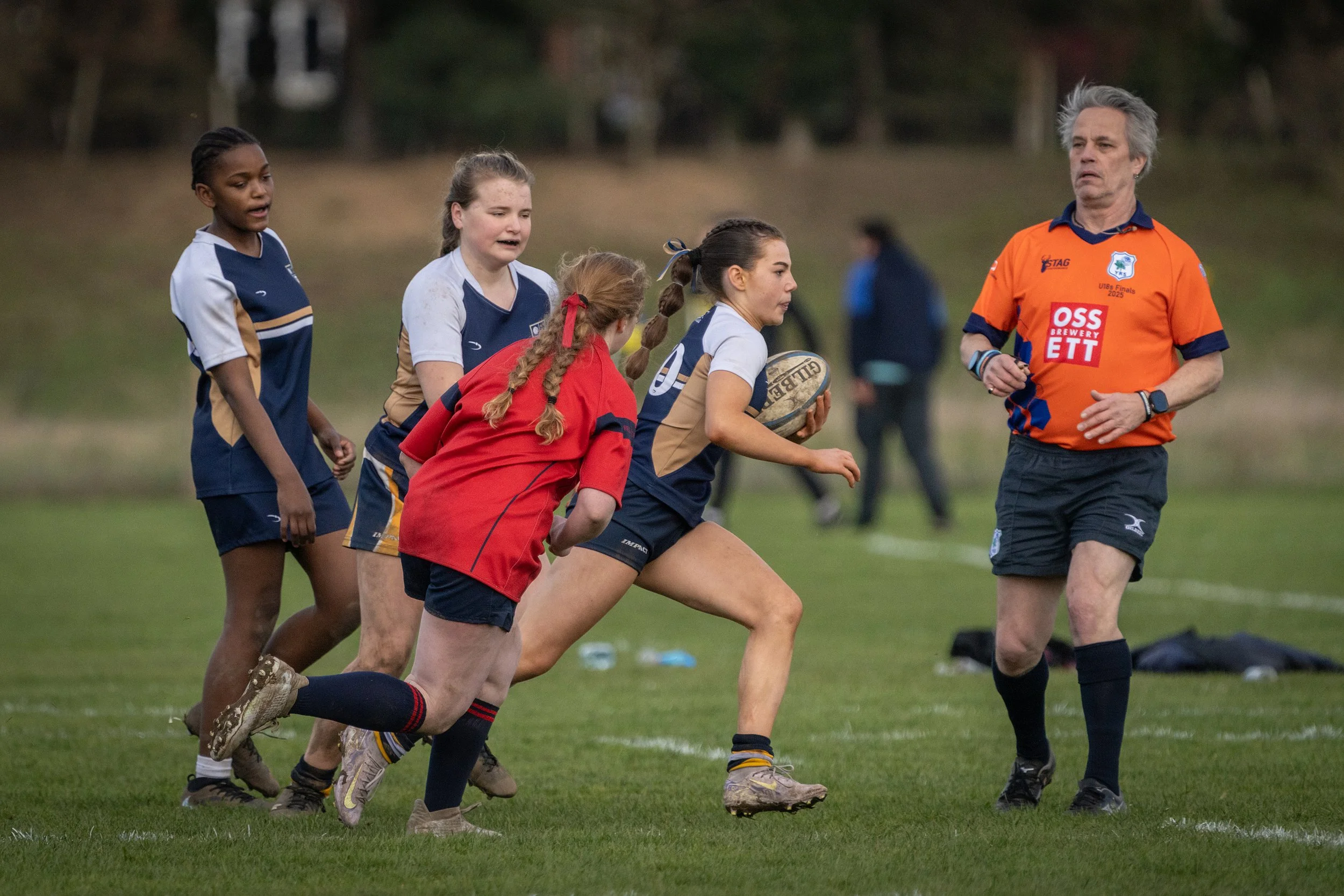 Young female rugby players running with a coach on a grassy field during a game, with trees in the background.
