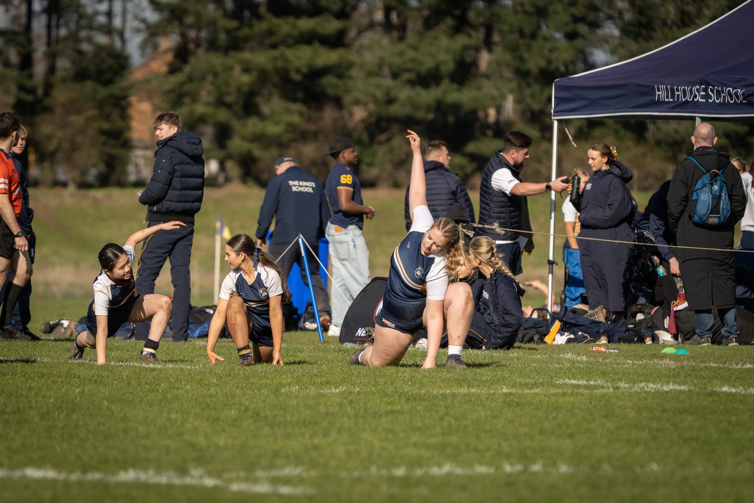 Students preparing for or participating in a sports event on a grassy field with a mix of people standing and kneeling, some under a blue canopy tent labeled 'Hillhouse School,' with trees in the background.