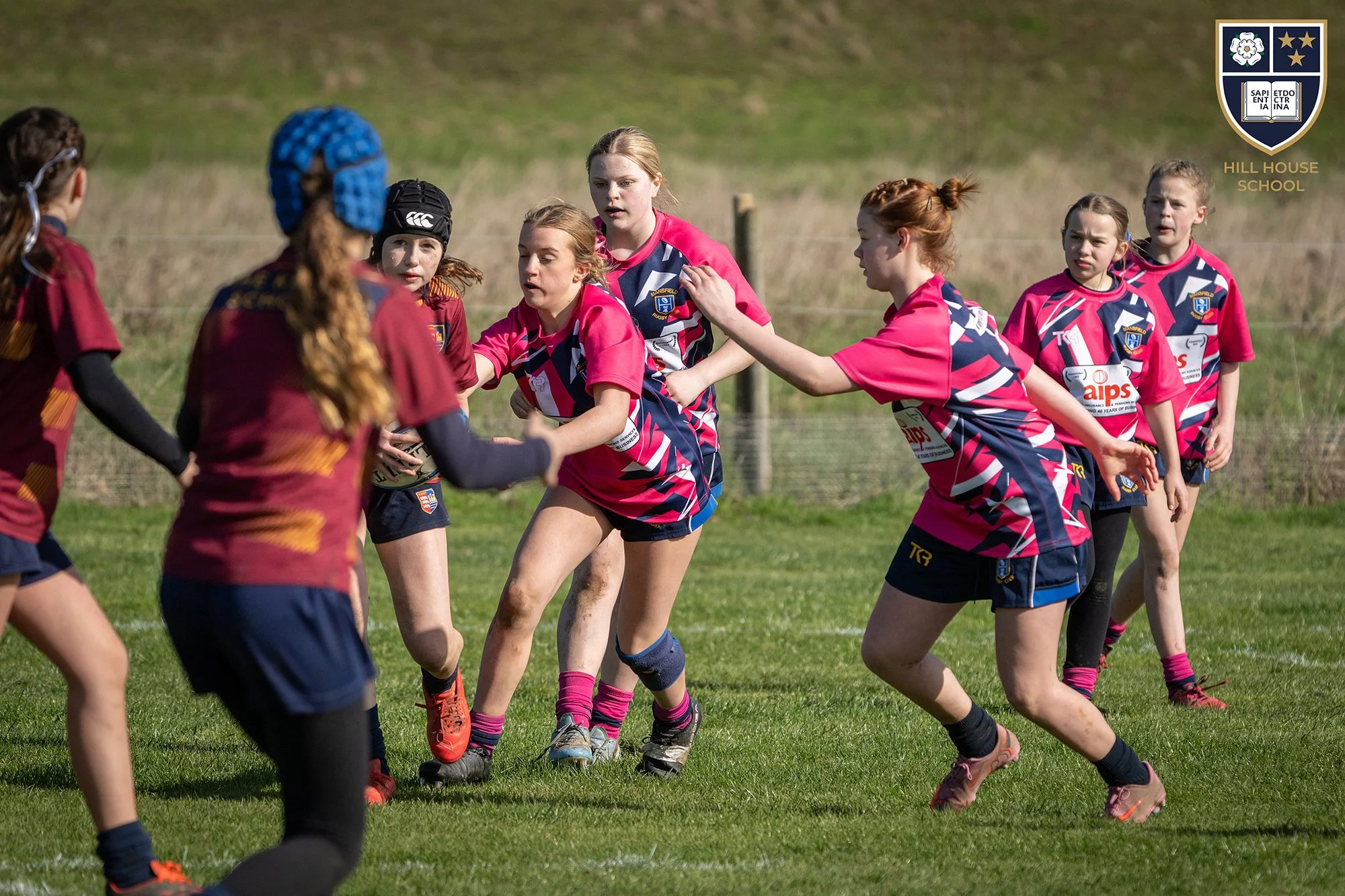 Young girls playing rugby on a grassy field during daytime, wearing pink and navy sports jerseys with a school crest, some with protective headgear, engaged in a game.