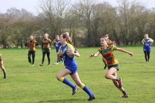 Girls playing rugby on a grassy field with trees in the background, some running with a rugby ball and others chasing.
