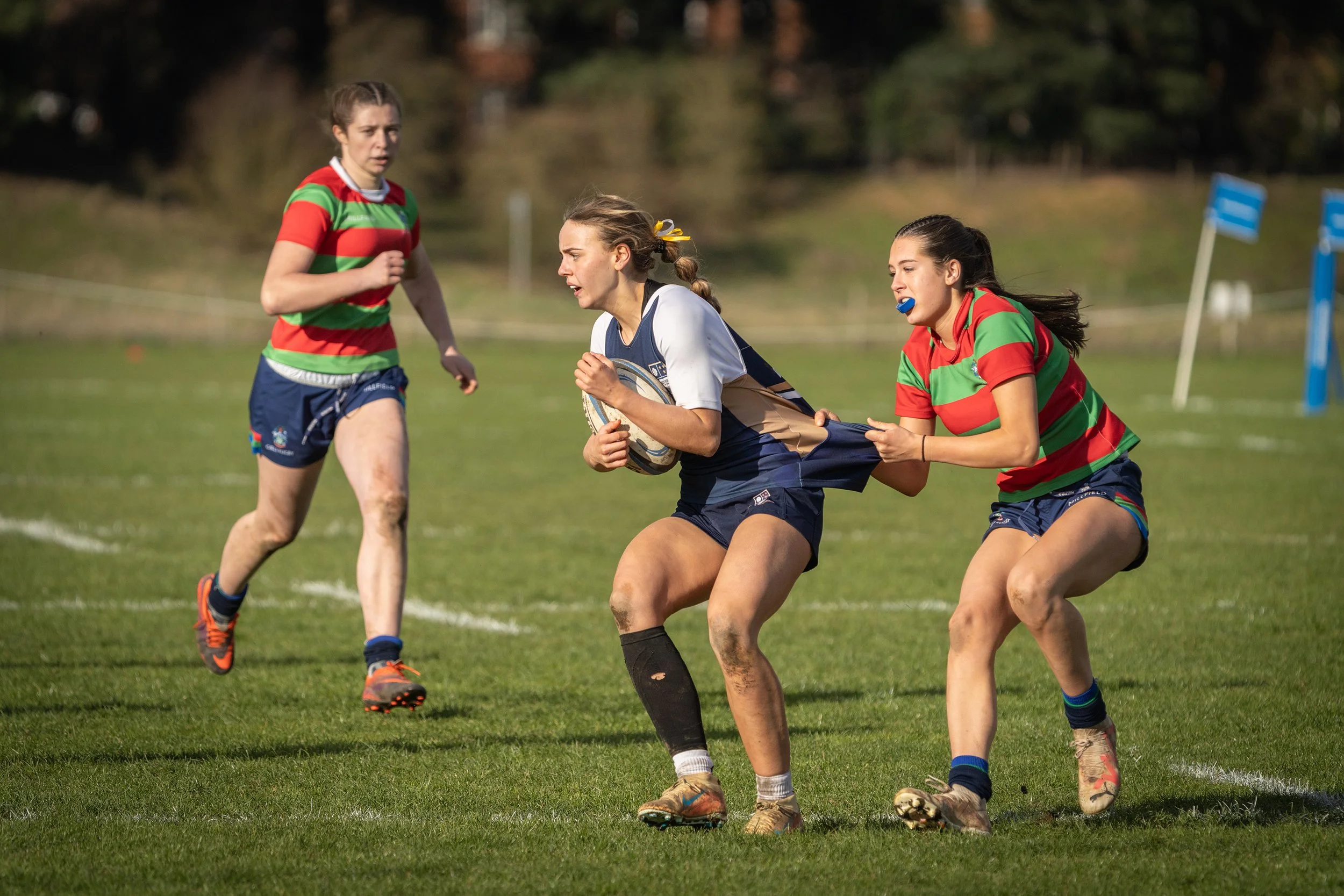 Three women playing rugby on a grassy field, with one woman running with the ball while two others attempt to tackle her.