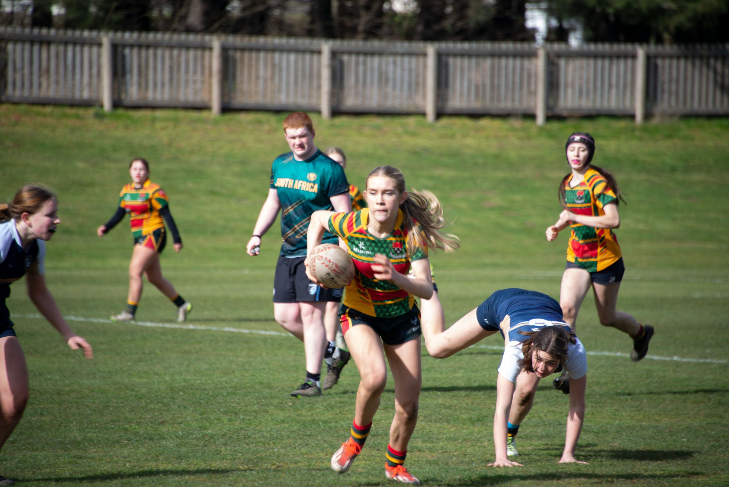 Women playing rugby on a grassy field, some running and tackling, with a wooden fence and trees in the background.
