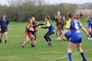 Girls playing rugby on a grassy field, wearing colorful uniforms, with some running and others tackling.