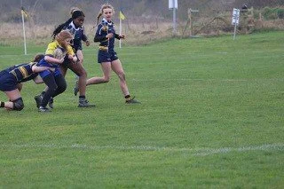 Youth girls playing rugby on a grassy field, with one girl running toward the try line while others are engaged in the game.