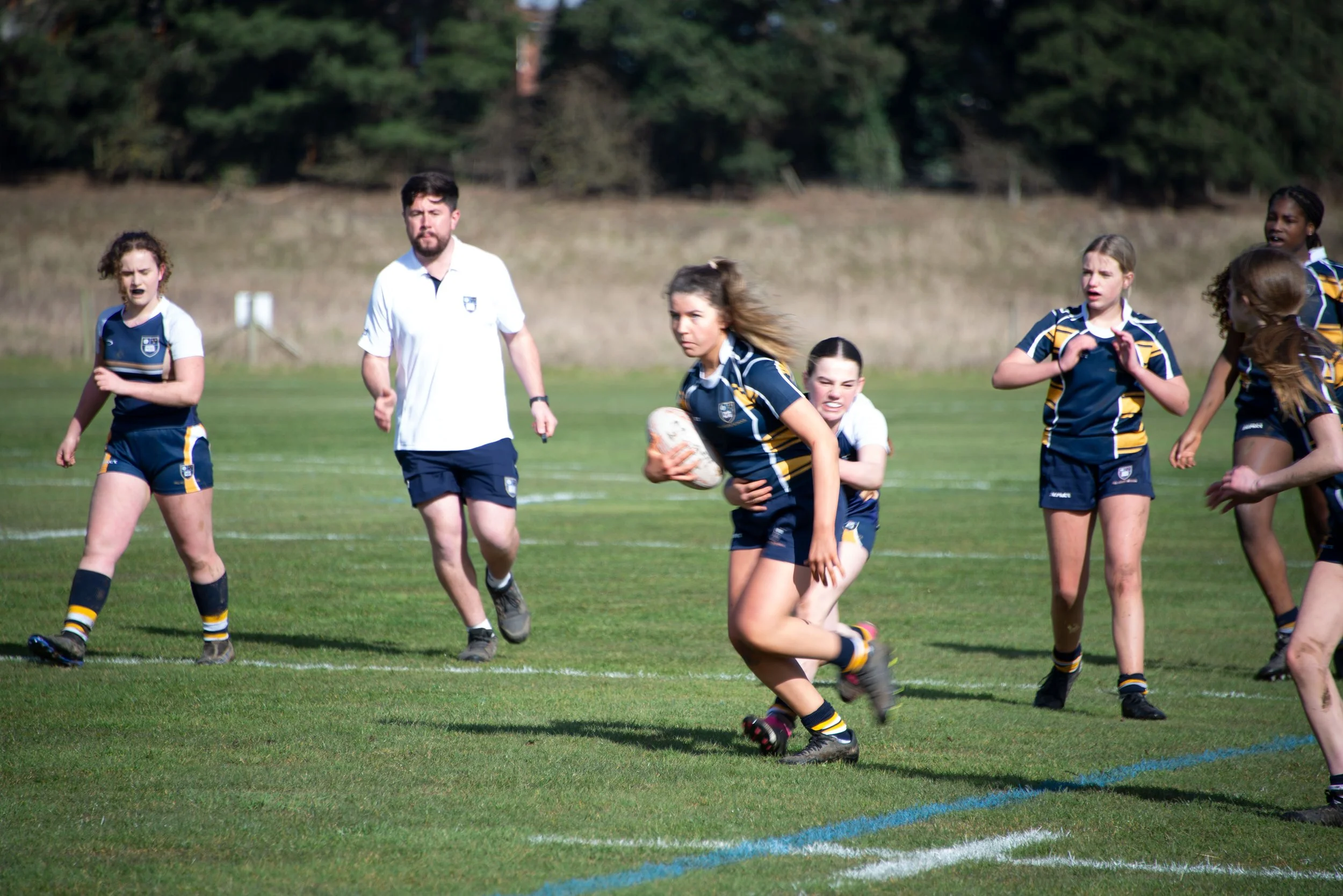 A girls' rugby team in blue and yellow uniforms playing on a grass field, with one girl holding a rugby ball and running, while teammates and a coach observe.