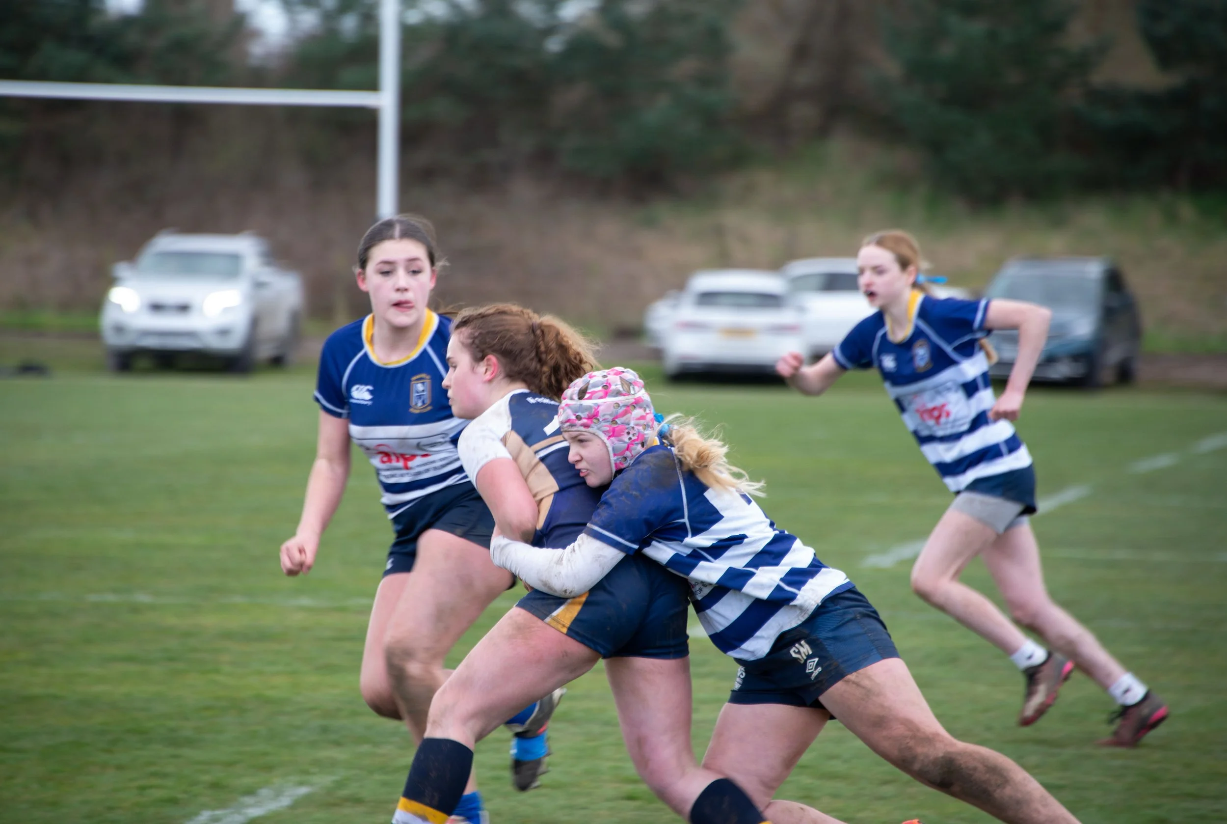 Girls playing rugby on a grassy field, wearing blue and white uniforms, with trees and parked cars in the background.