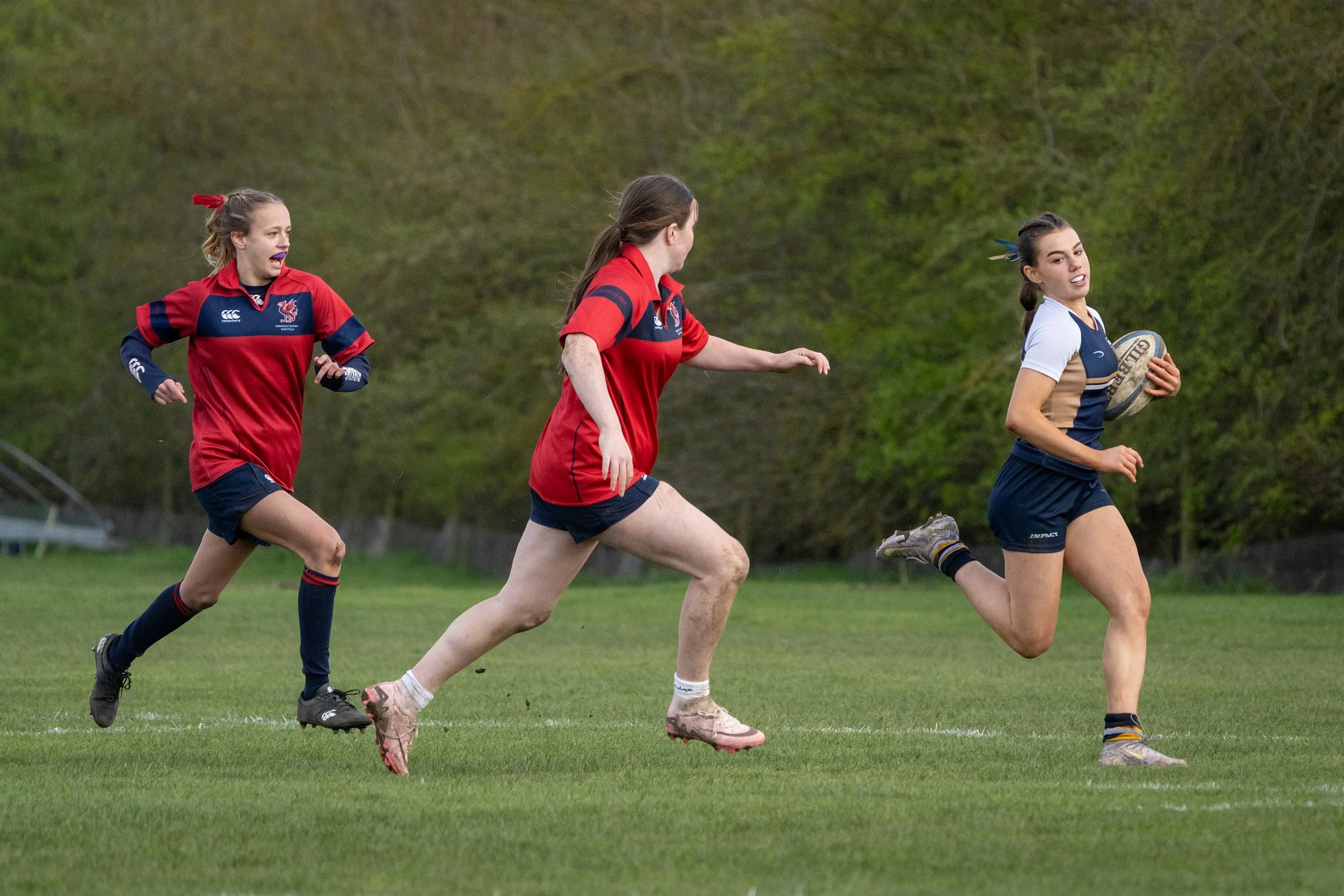 A group of young women playing rugby on a grassy field, wearing sports uniforms, with a background of trees.