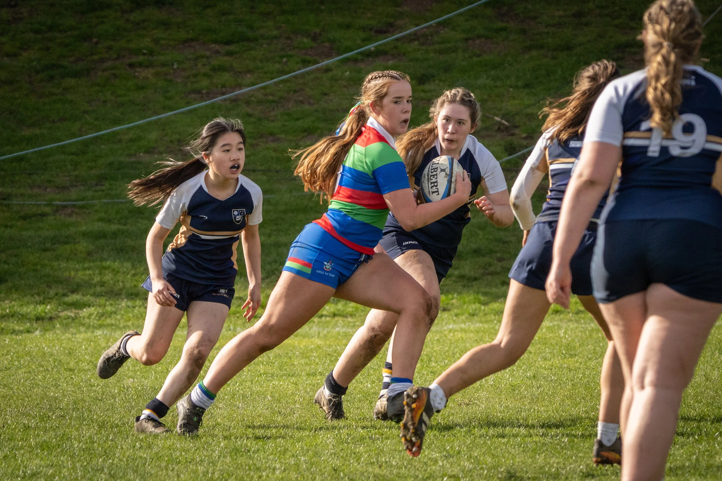 Girls playing rugby on a grassy field, one girl in a rainbow-colored jersey holding a rugby ball, others running after her.